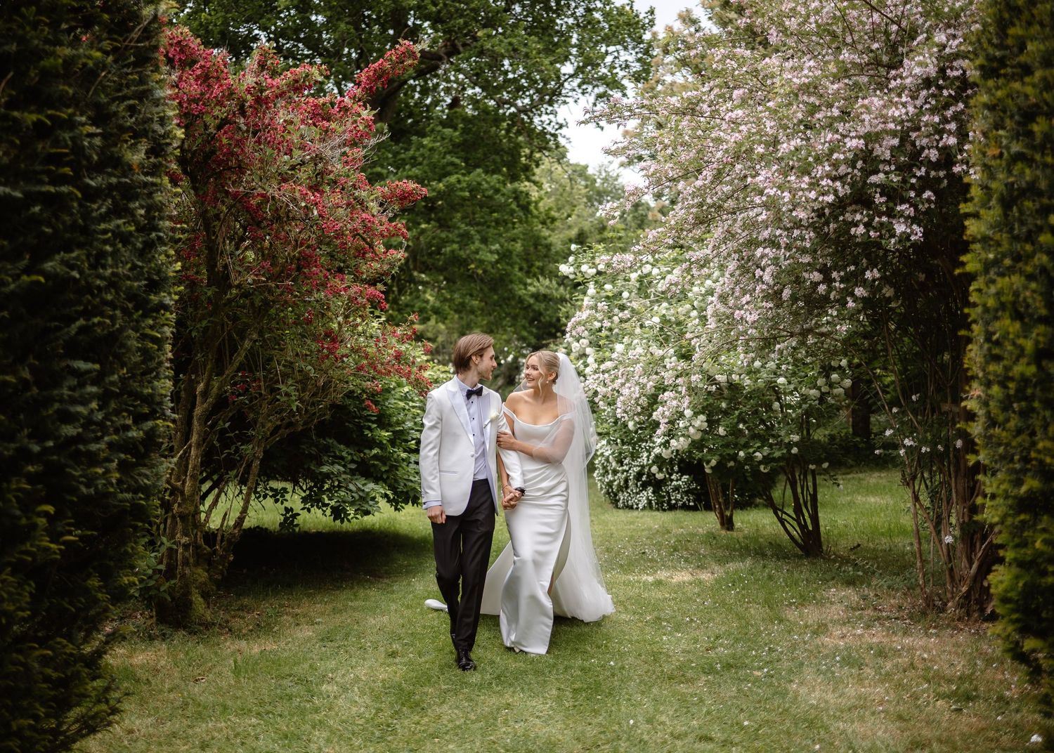 Bride and groom stroll through a garden path framed by blooming red and white flowering trees on wedding day.