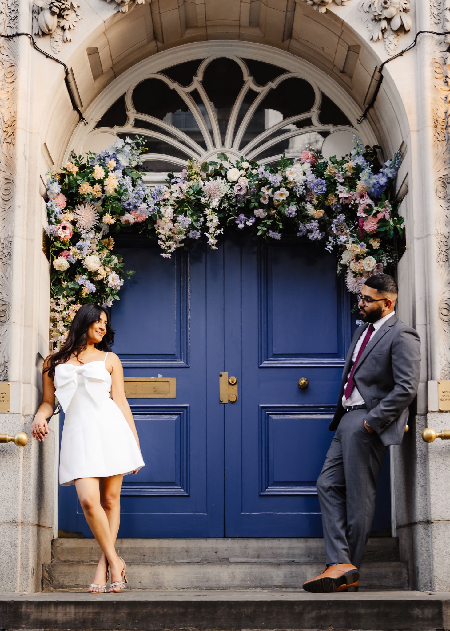 Couple posing in front of elegant blue door adorned with colorful floral arch at historic building.