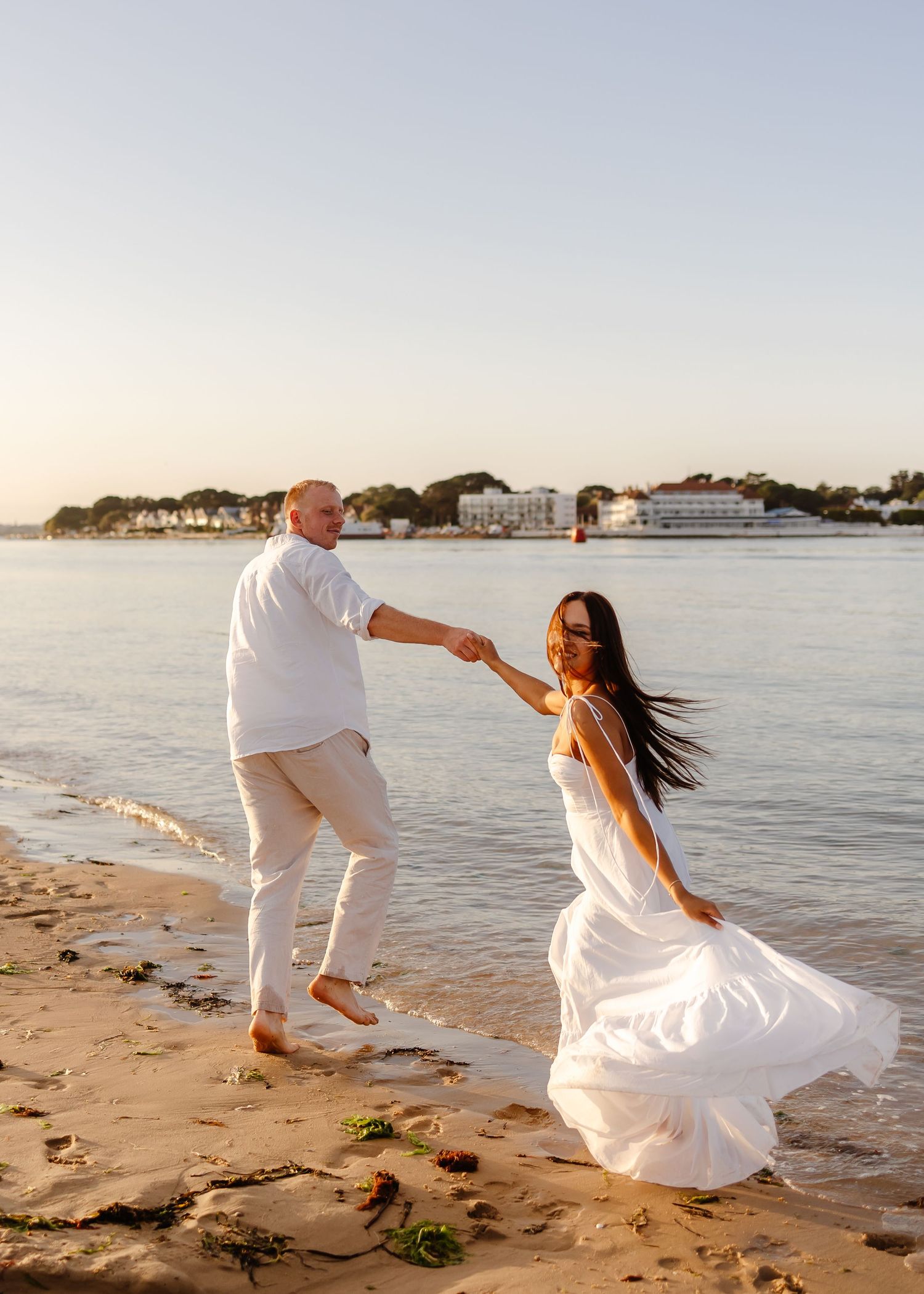 Couple in white outfits holding hands on a sandy beach at golden hour with waterfront buildings in the background.