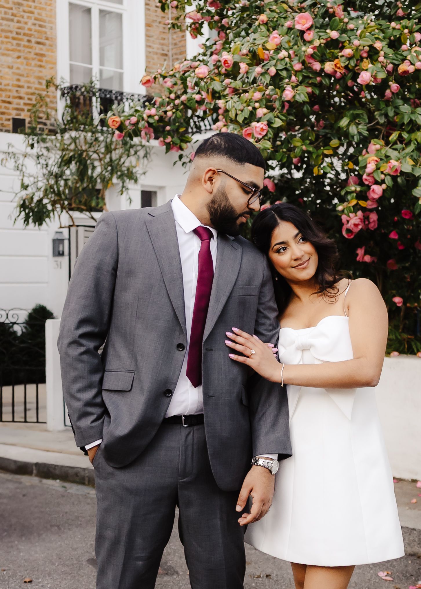 A couple in wedding attire poses affectionately against a brick wall with pink flowering bushes in multiple shots.