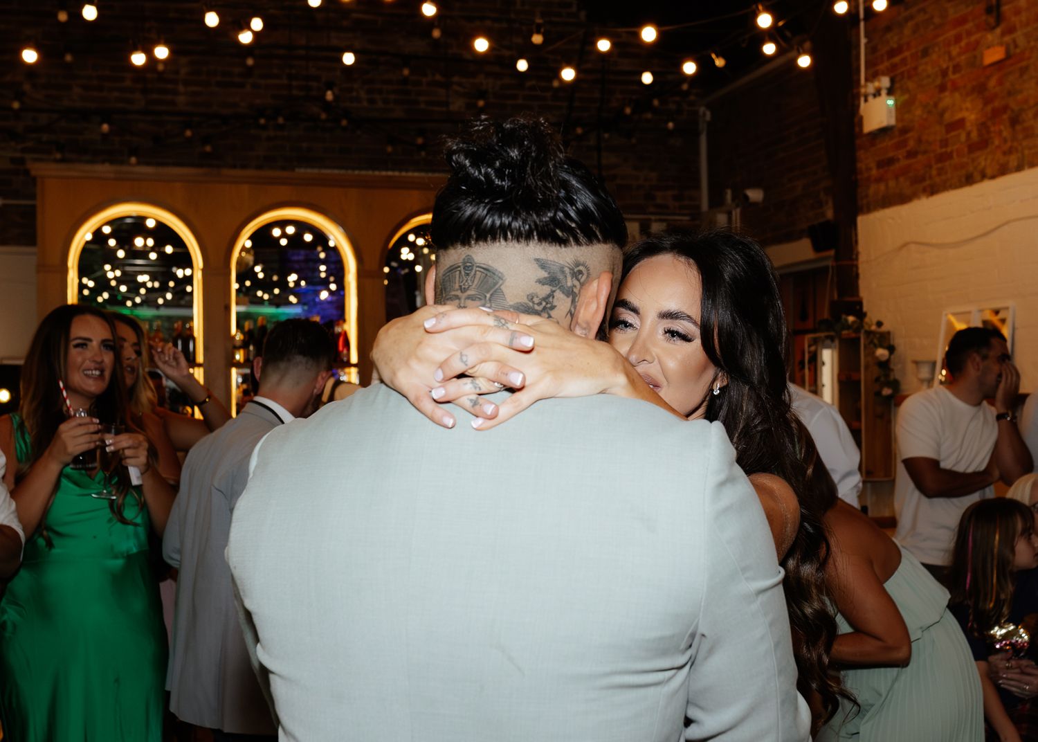 A couple embraces on the dance floor at a wedding reception with string lights and arched windows in the background.