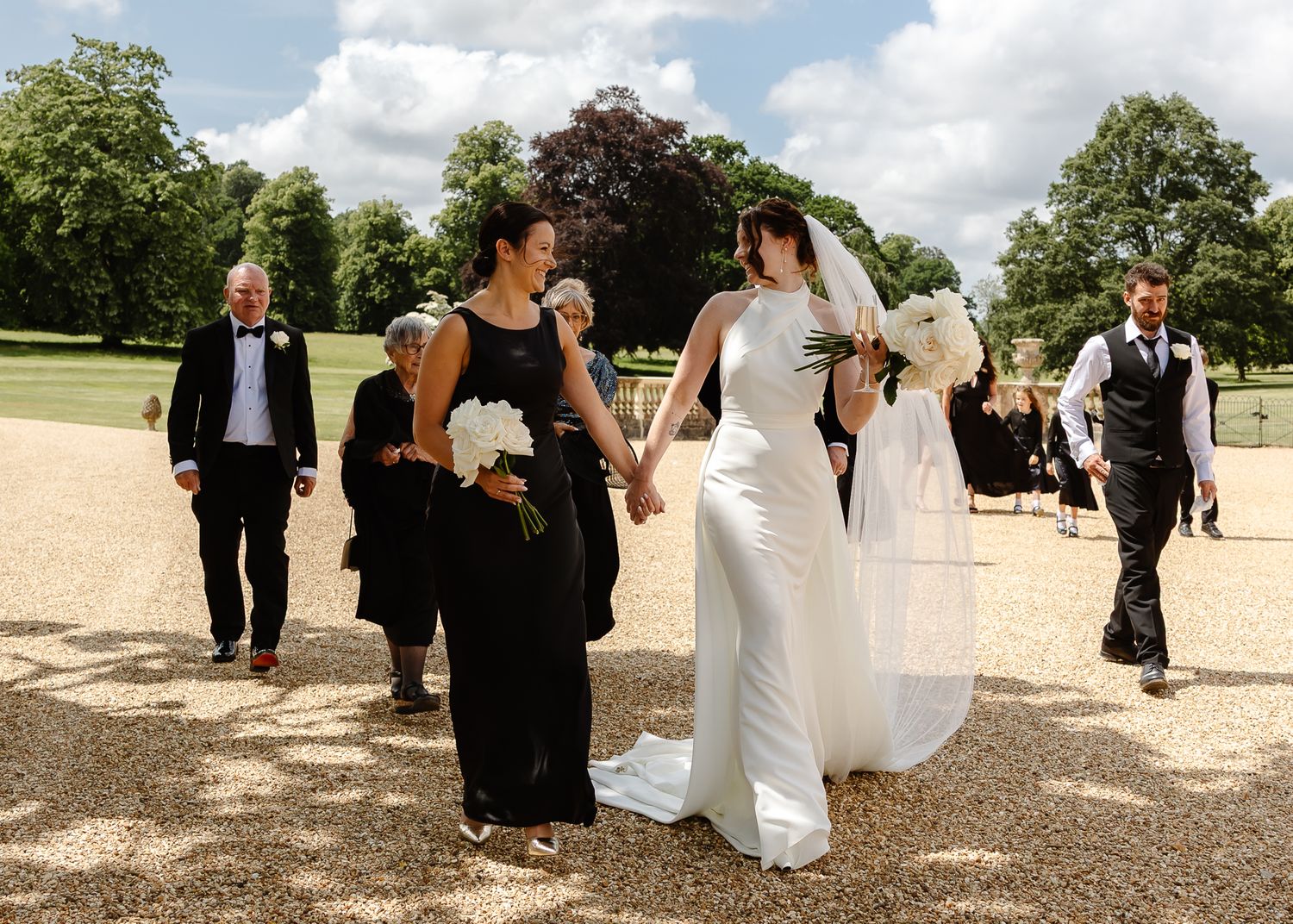 Wedding celebration on gravel path with guests in formal attire walking through garden grounds.
