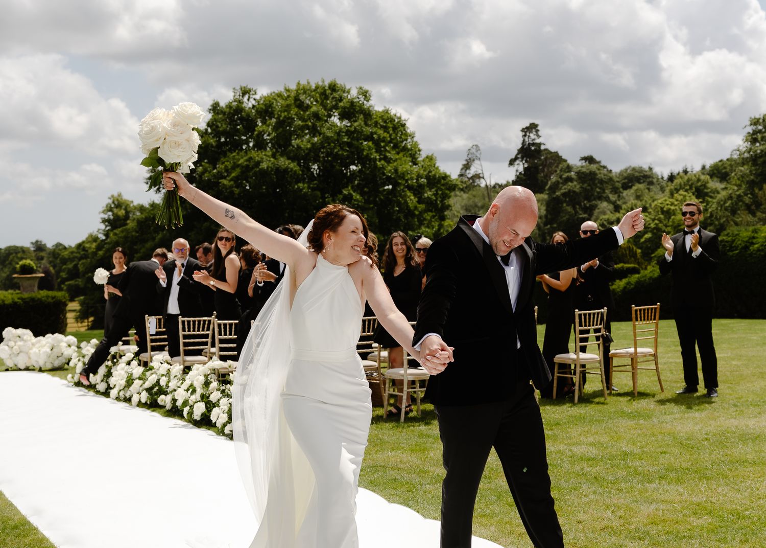 Wedding party members celebrate joyfully in a grassy outdoor setting with their arms raised in celebration.