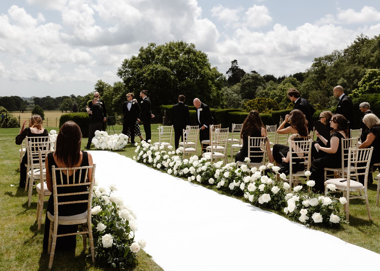 An outdoor wedding ceremony setup with white floral arrangements lining the aisle and guests seated in wooden chairs.
