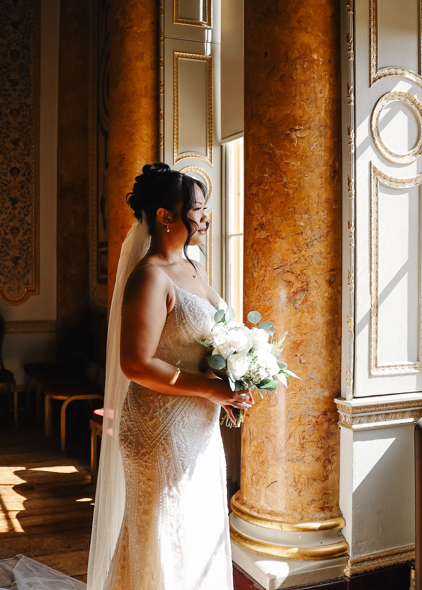 A bride in a white dress stands near ornate marble columns in an elegant room with natural window light.