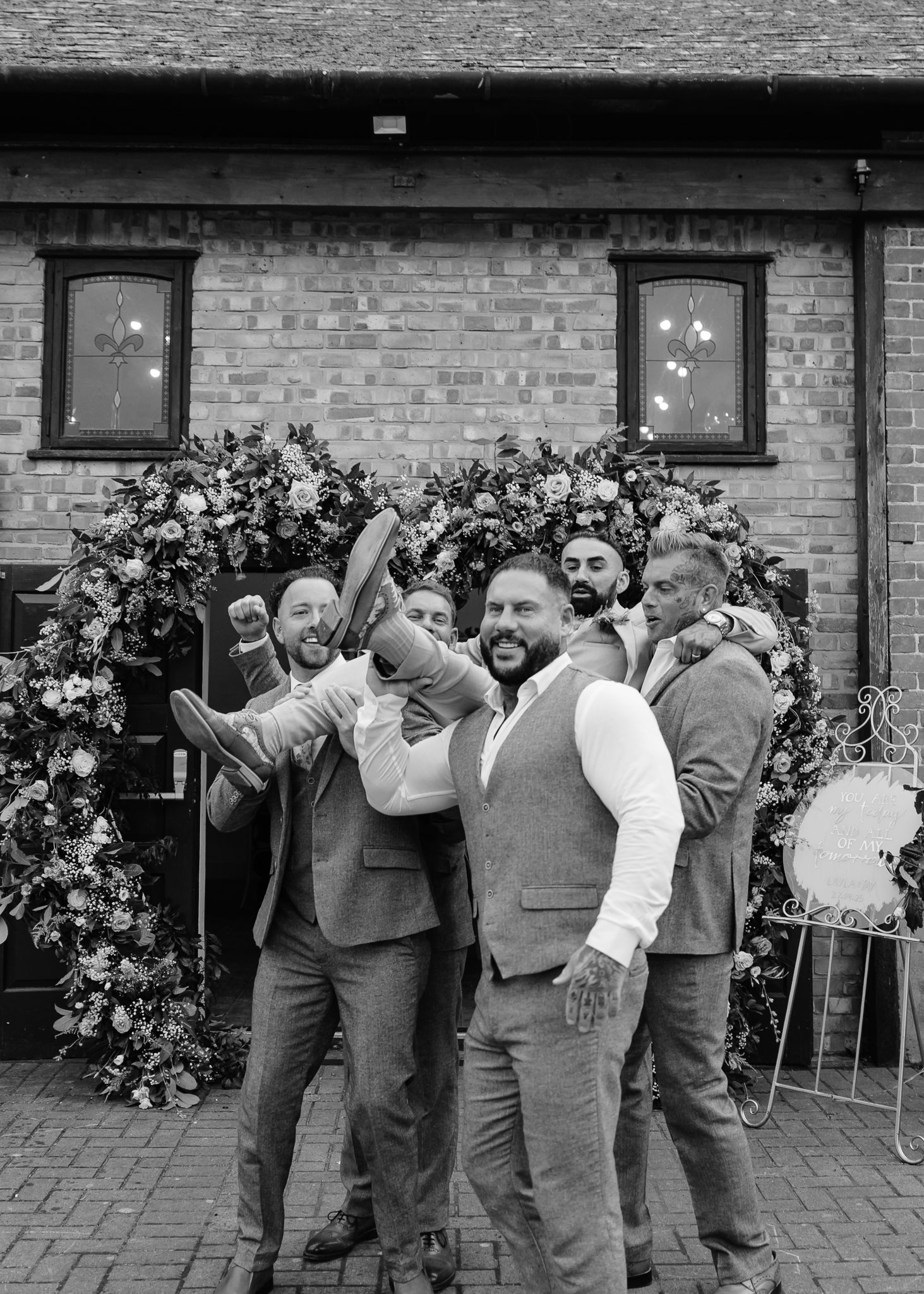 Wedding party poses playfully for group photos against a brick wall with floral decorations.