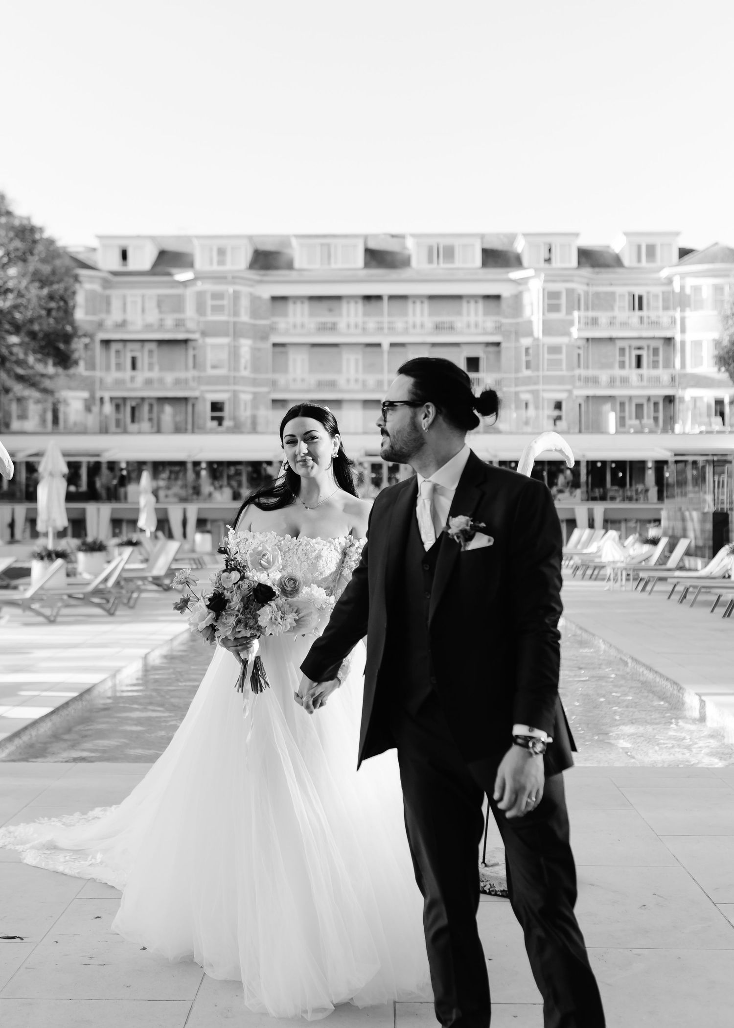 Black and white photo of a bride and groom holding hands near a pool with a grand hotel building in the background.