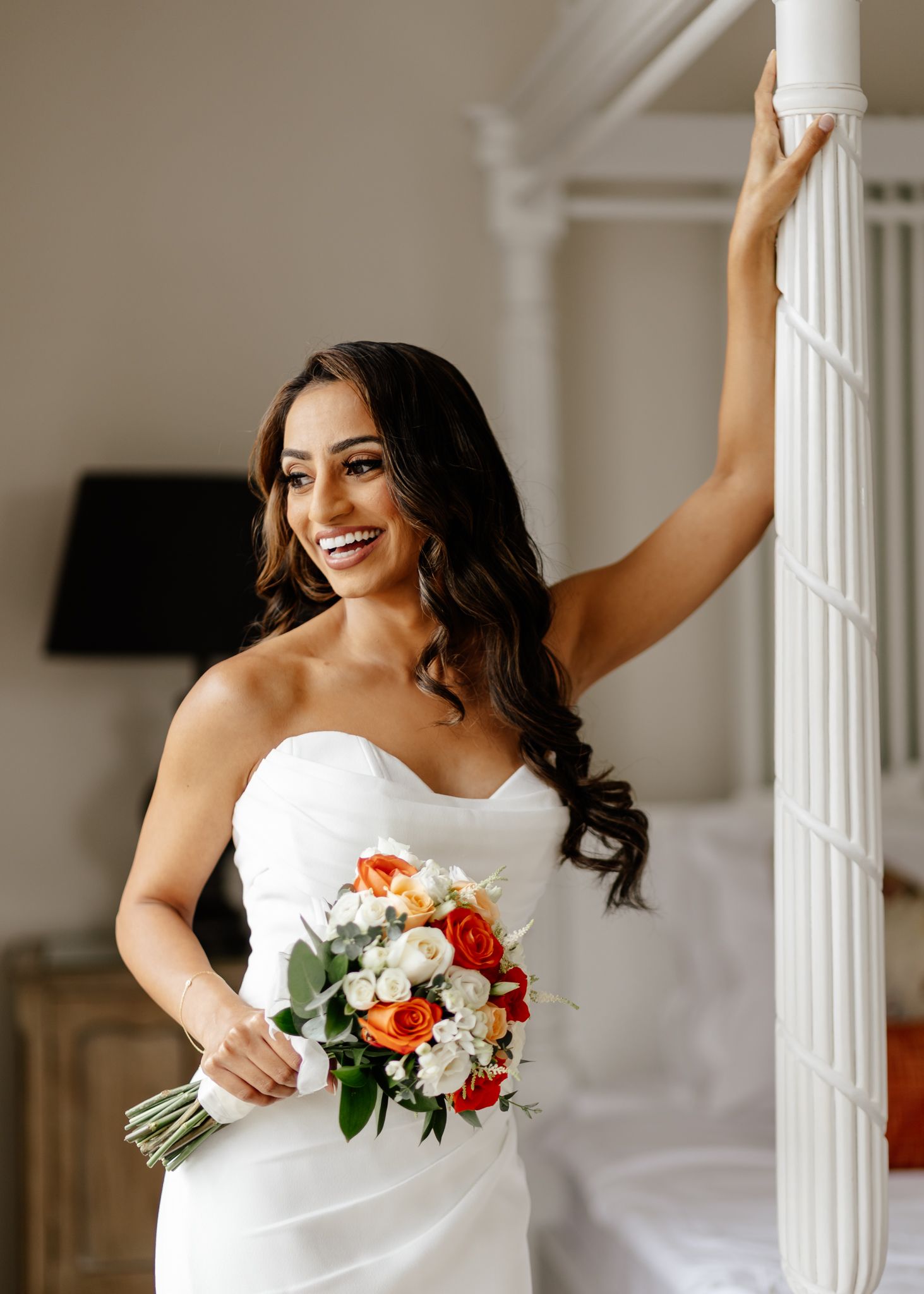 A bride in a white strapless wedding dress holds a bouquet of red and white flowers against a light interior backdrop.