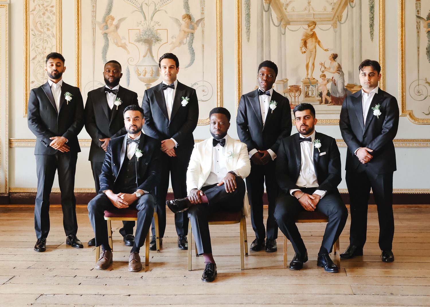 Groomsmen in formal attire sitting in a row against an ornate wall in an elegant venue.