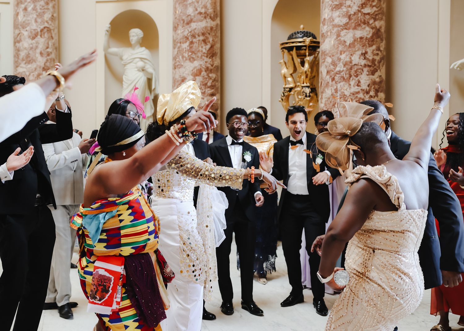 Wedding reception guests celebrate and dance in ornate ballroom with classical murals and columns.