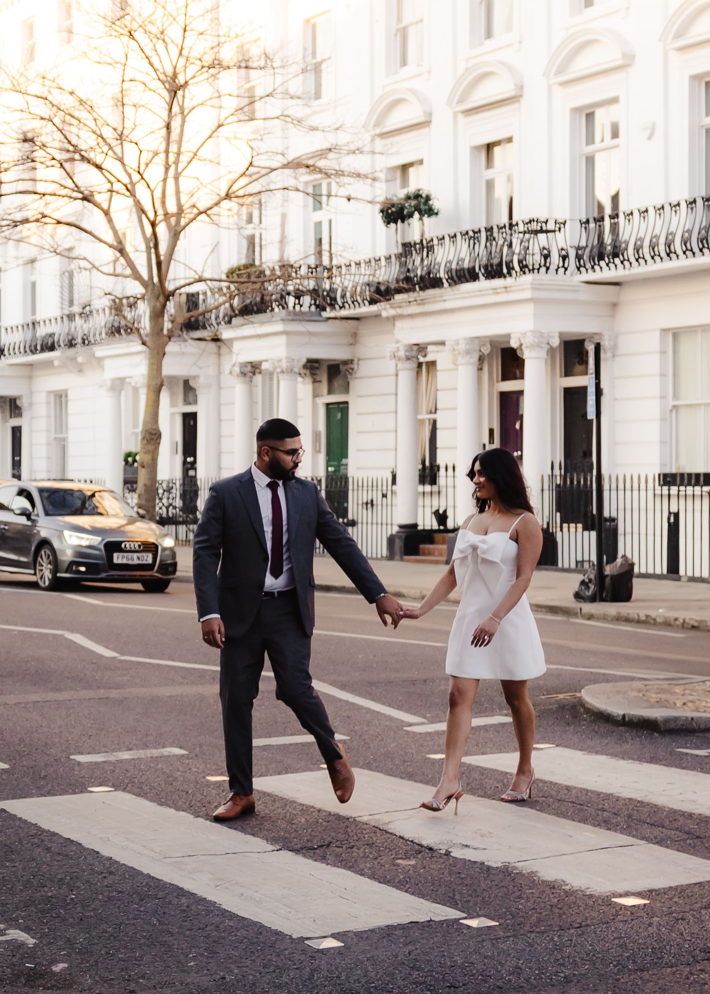A couple in formal attire holds hands while crossing a street in front of elegant white townhouses in London.