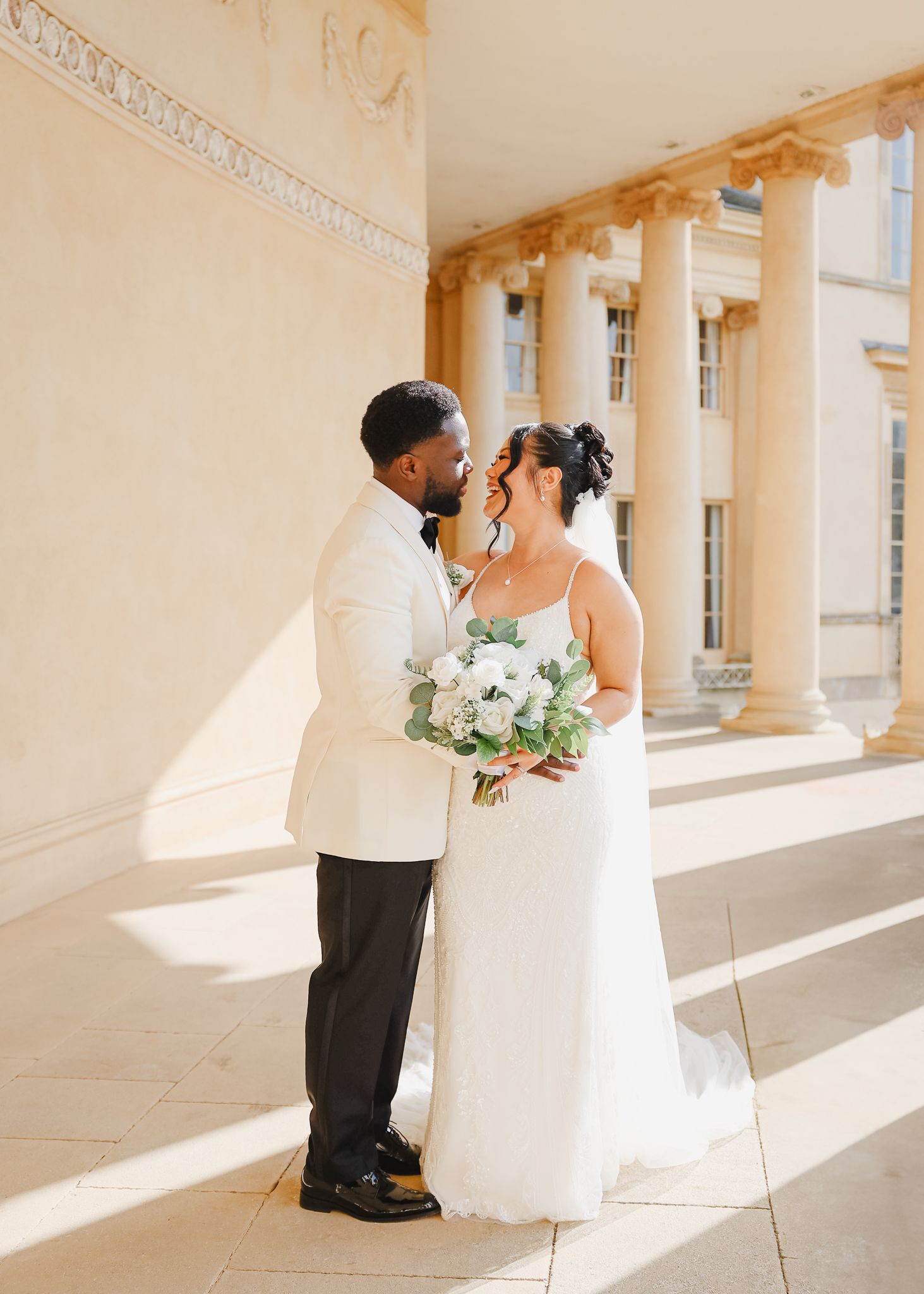 A romantic wedding couple embraces in a sunlit colonnade with elegant white columns casting dramatic shadows.