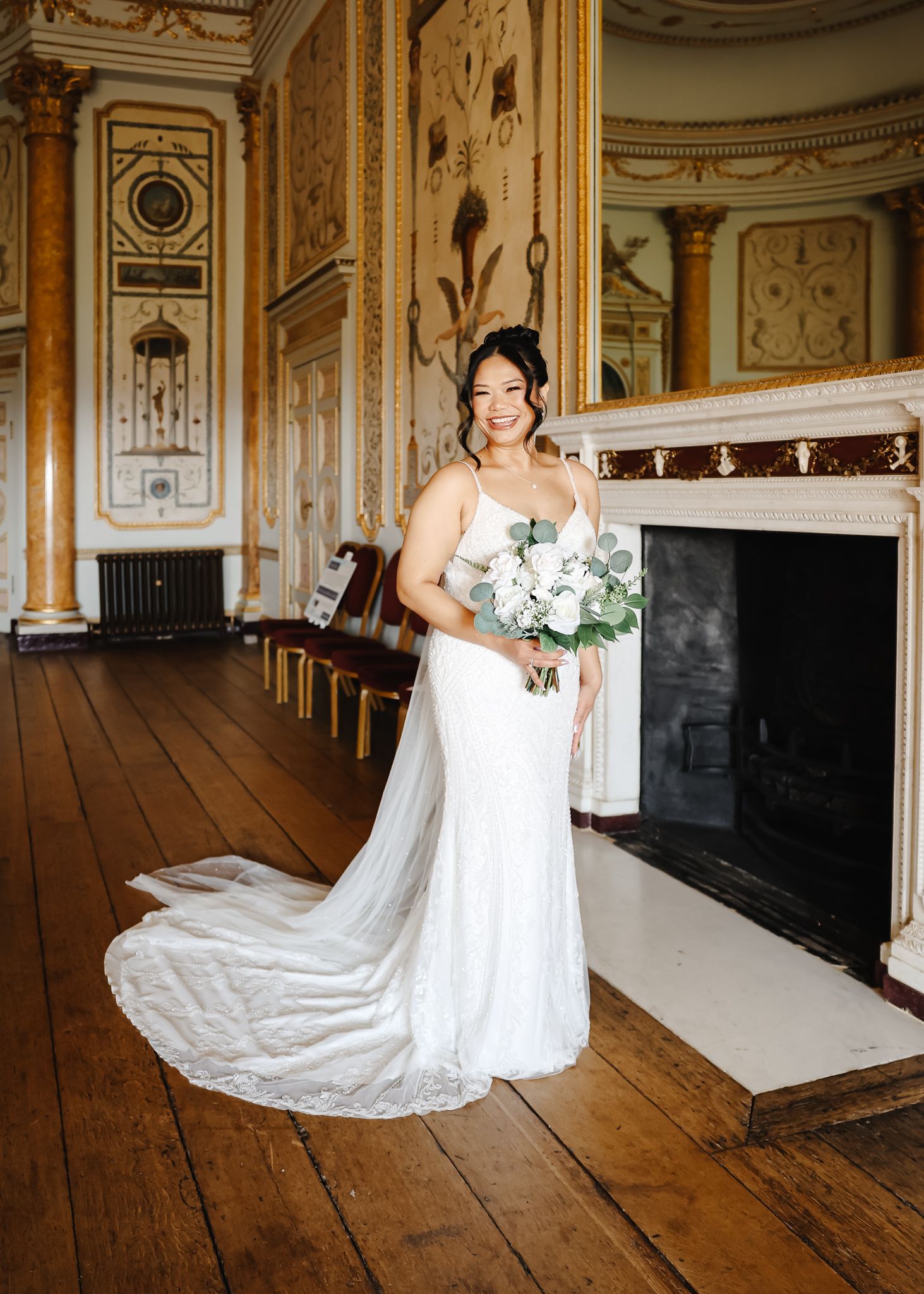 A bride in a white lace wedding gown poses in a grand historic room with ornate walls and wooden floors.