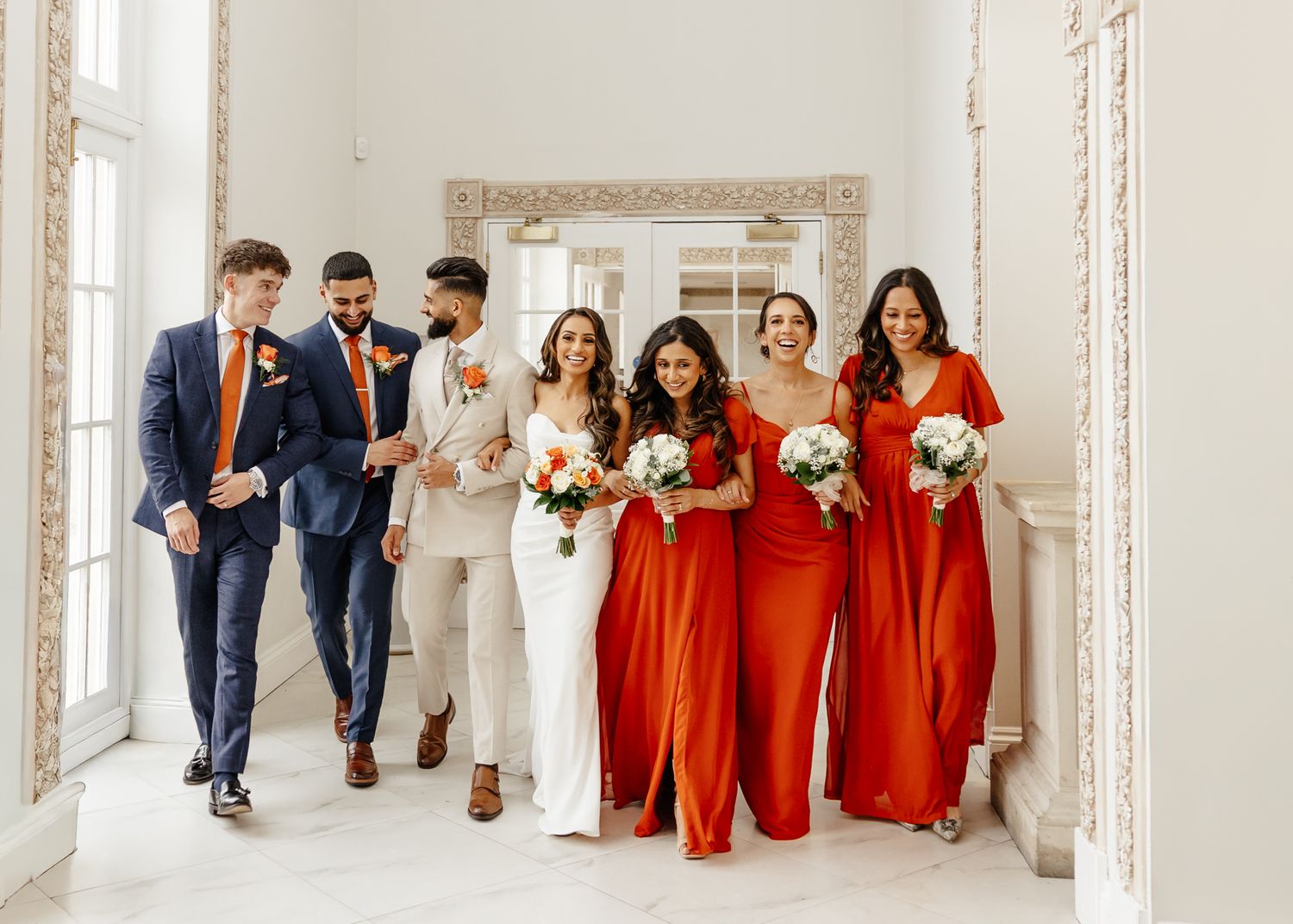 Wedding party in formal attire walks and poses together in an elegant white hallway with ornate mirrors.