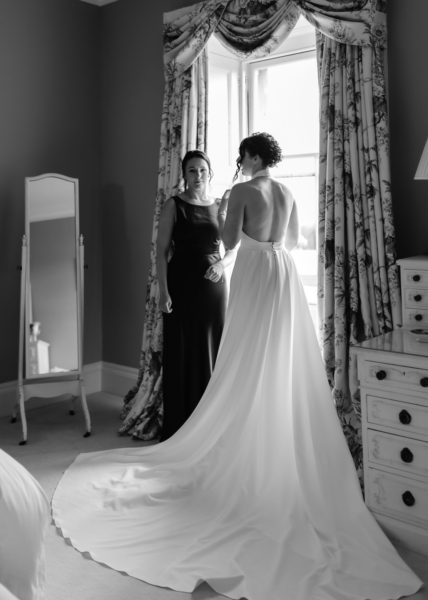 A black and white photo shows a wedding dress fitting in an elegant room with ornate curtains and furniture.