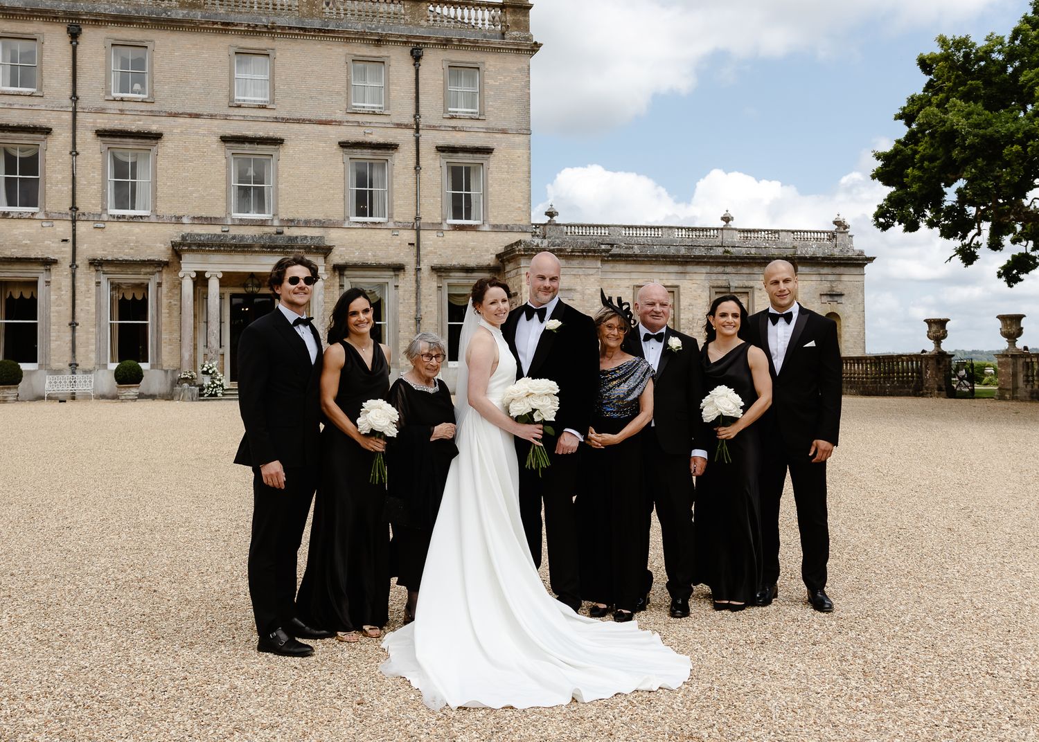 A wedding party poses together in formal attire outside a stately manor house.