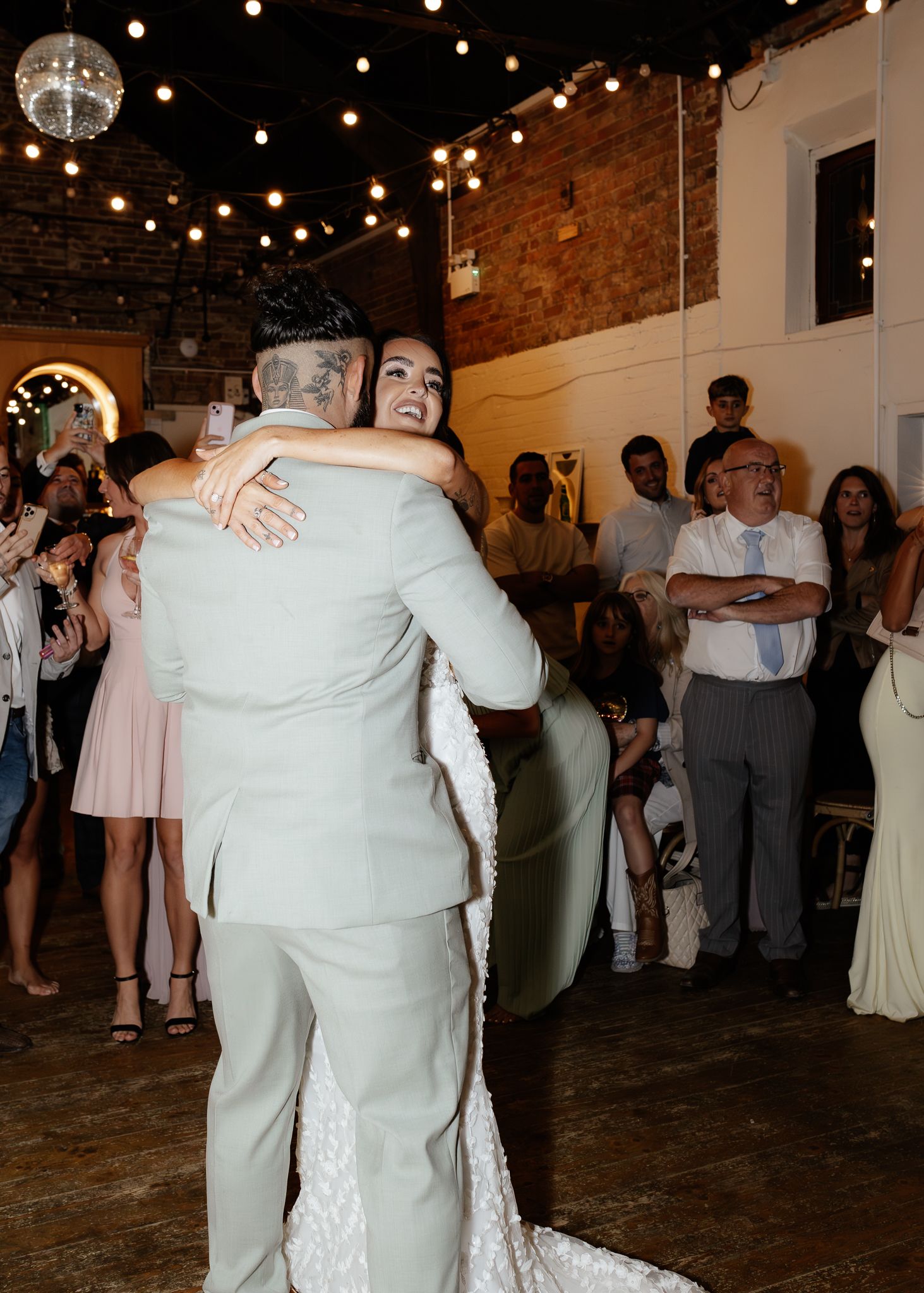 Two people share an emotional embrace on a dance floor at a wedding reception with string lights and exposed brick walls.