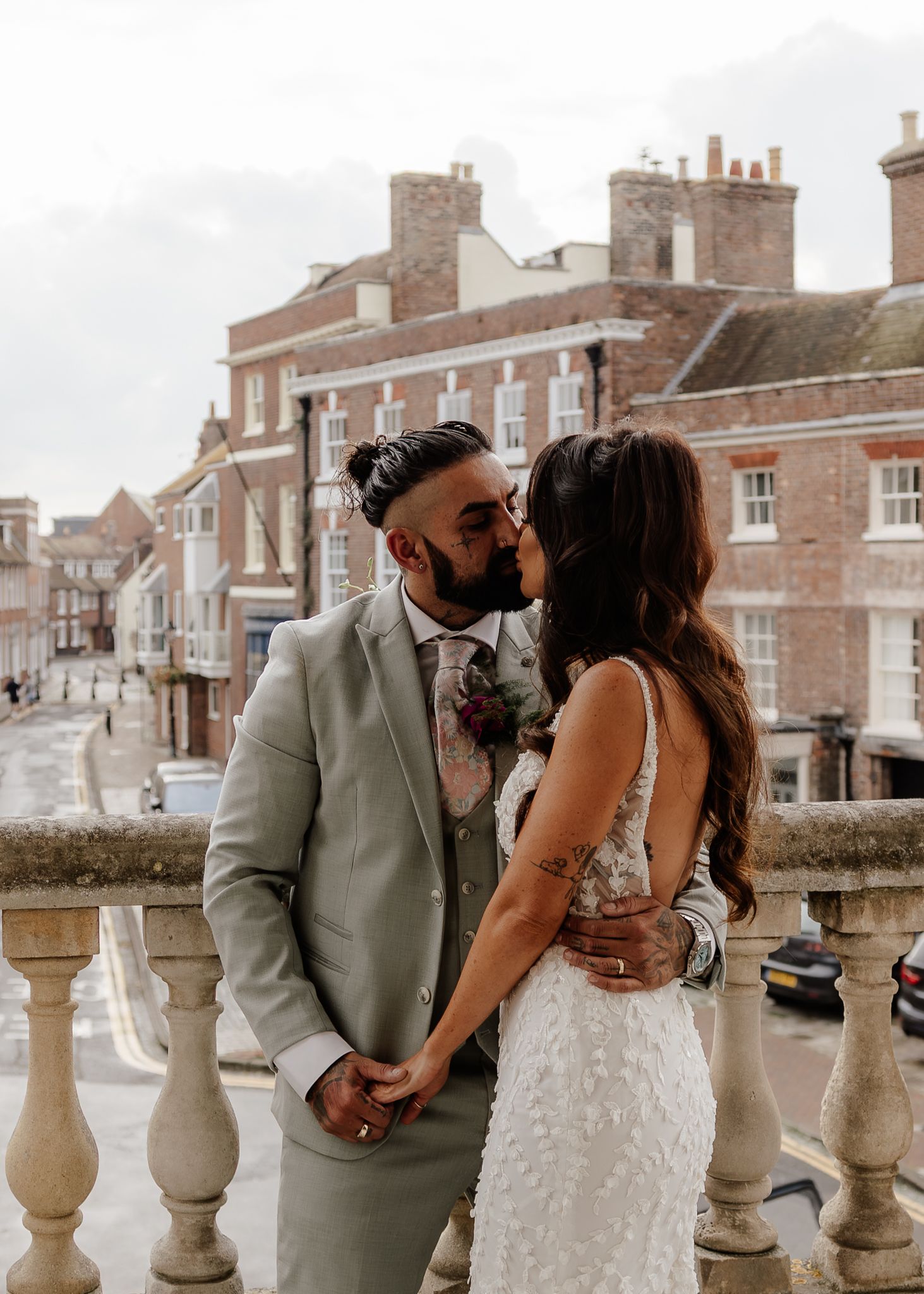 A romantic wedding couple share an intimate moment on a stone balcony overlooking a historic English town street.