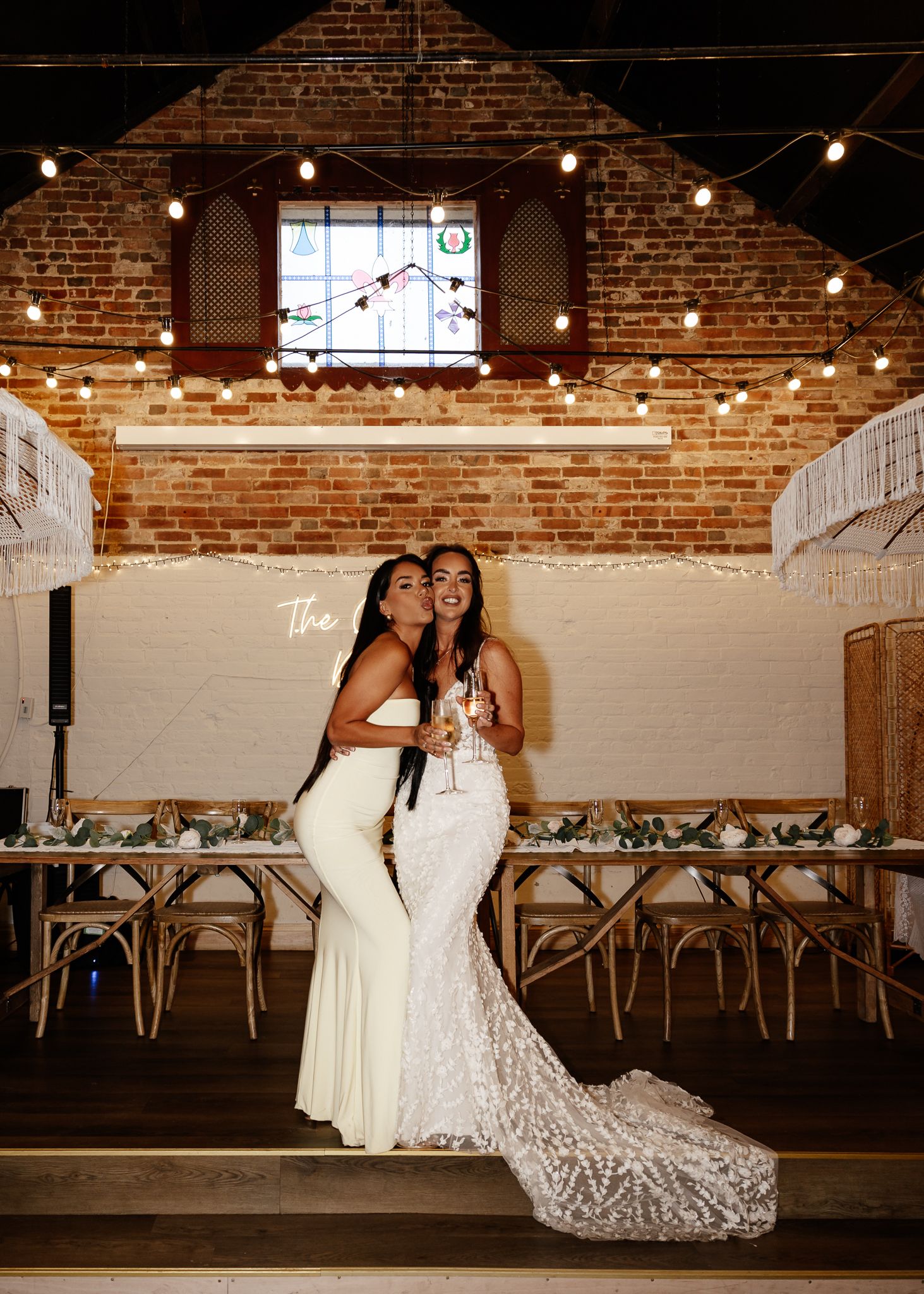 Two people in elegant white dresses pose together at a rustic brick venue with string lights and wooden chairs.