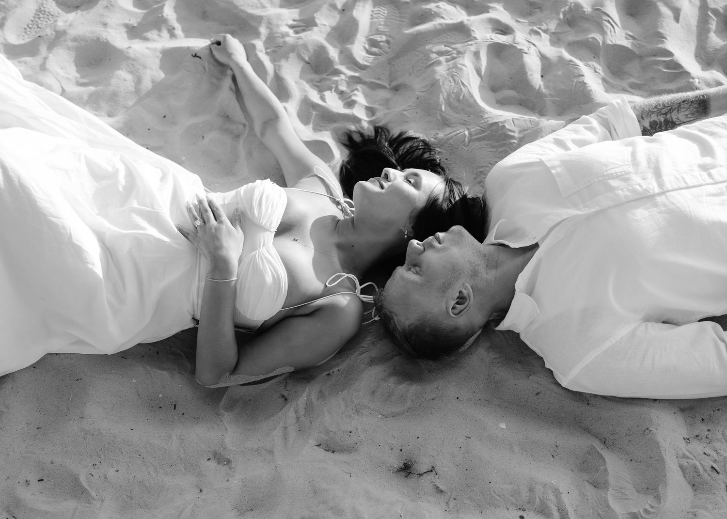 Black and white photo of couple lying on sandy beach with waves, wrapped in white fabric.