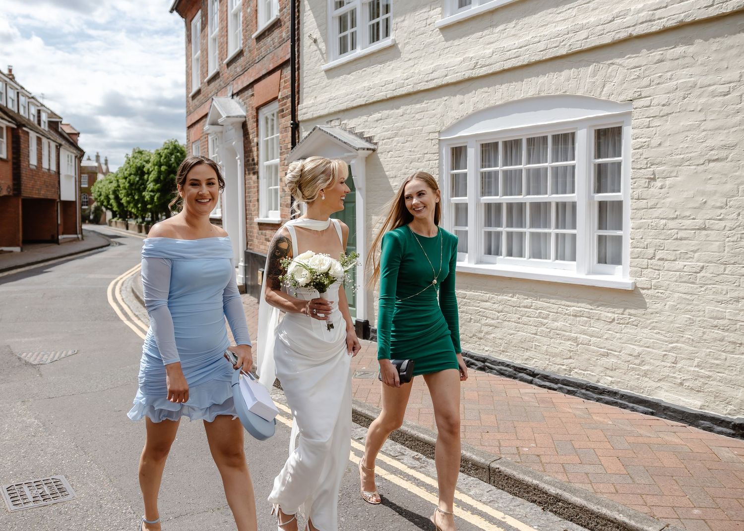 Bride in white gown walks with bridesmaids in blue and green dresses along a charming English street.
