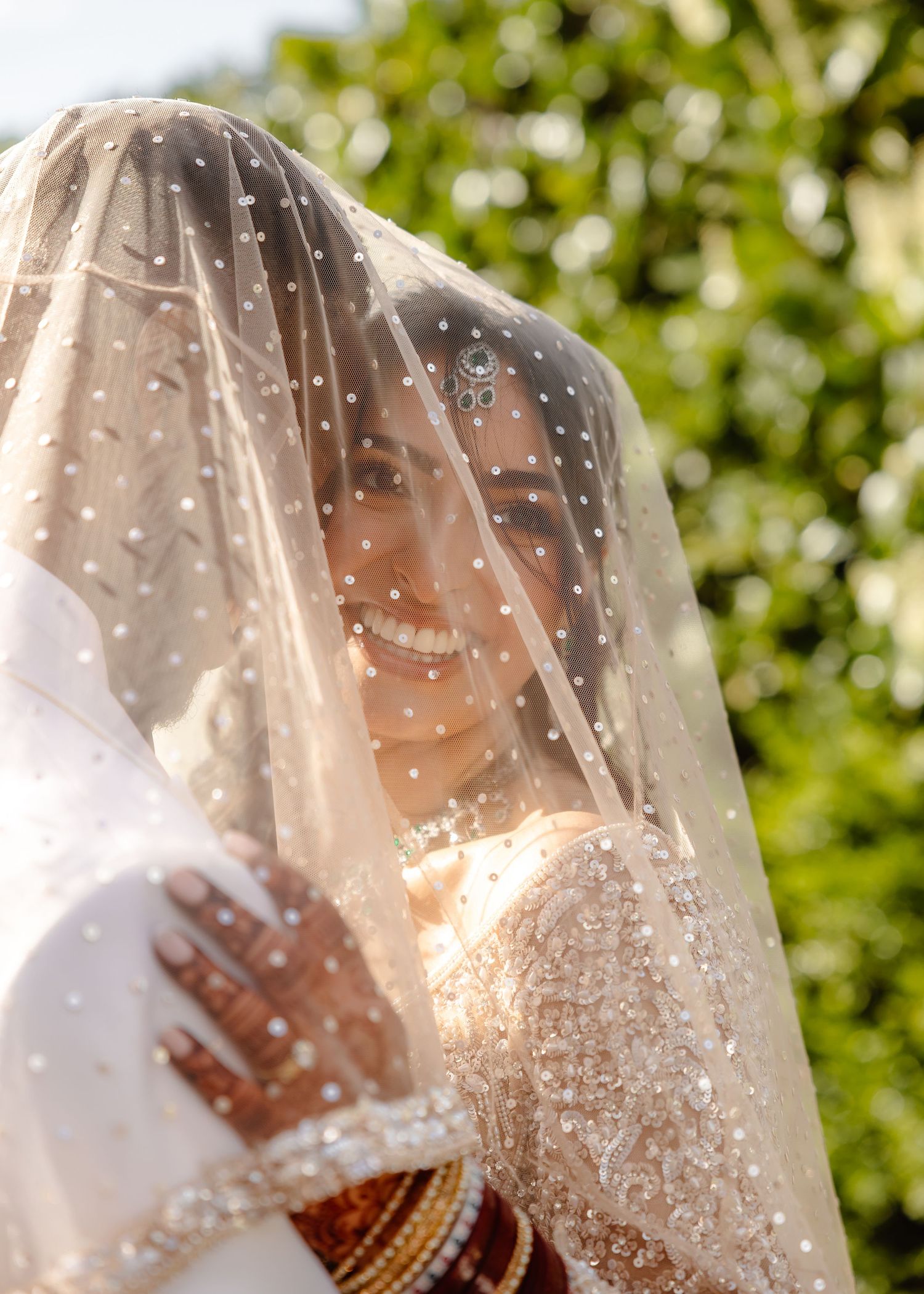Smiling bride wearing a pearl-studded veil and gold bangles at an outdoor wedding ceremony.