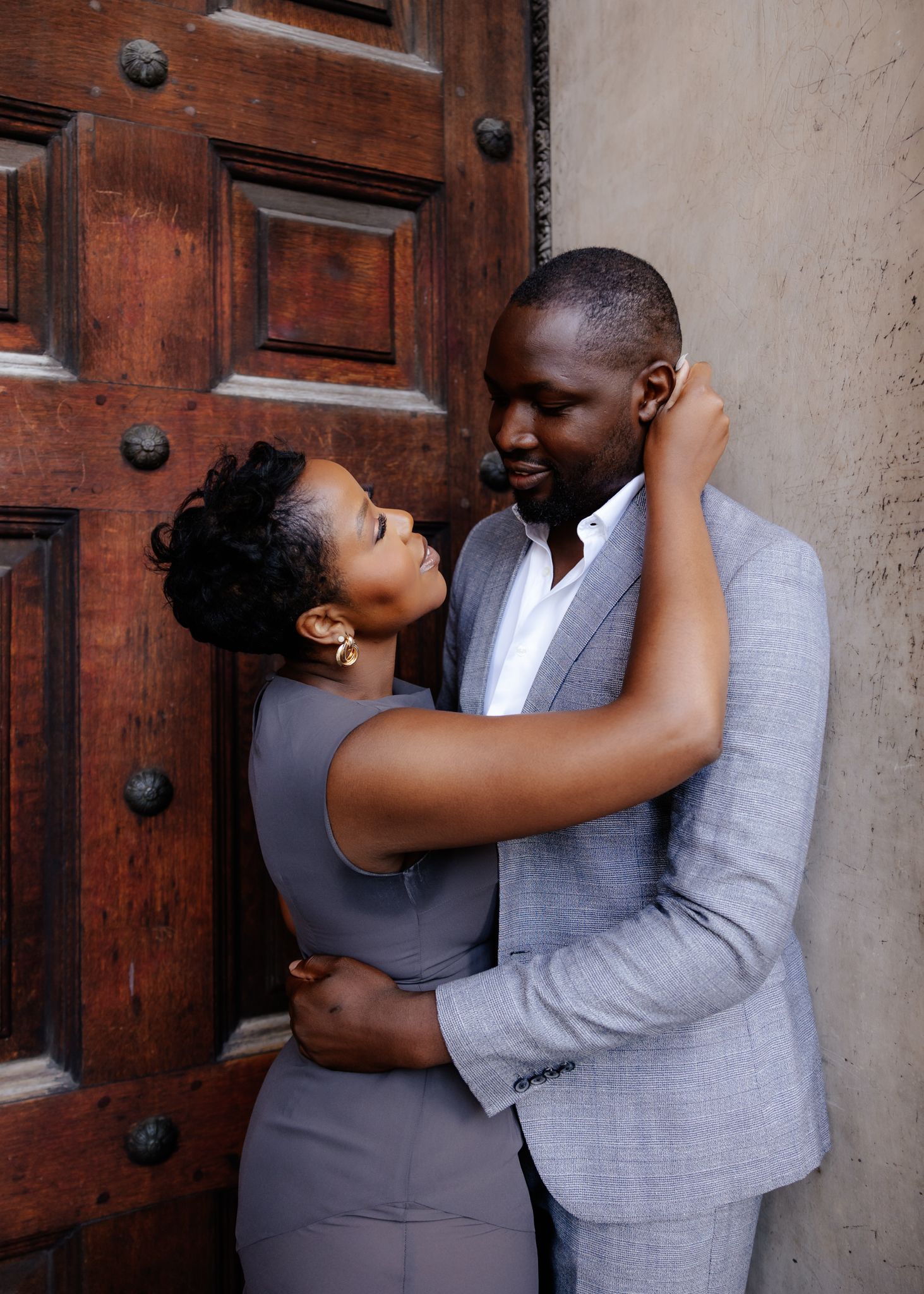 A couple in gray and navy outfits share an intimate moment in front of a rustic wooden door.