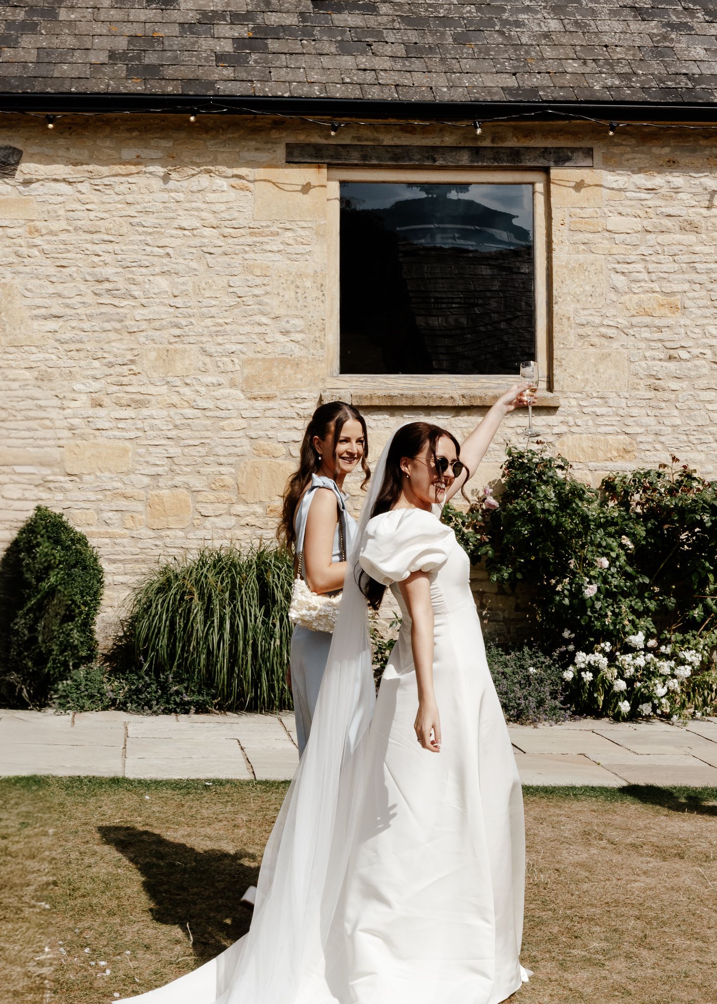 Two people in white dresses share a joyful moment together outside a stone building during a wedding celebration.