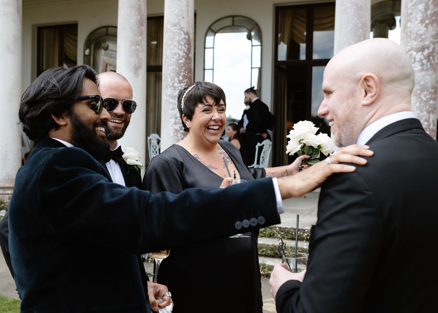 Wedding party members in formal attire gather and interact on a patio near columns.