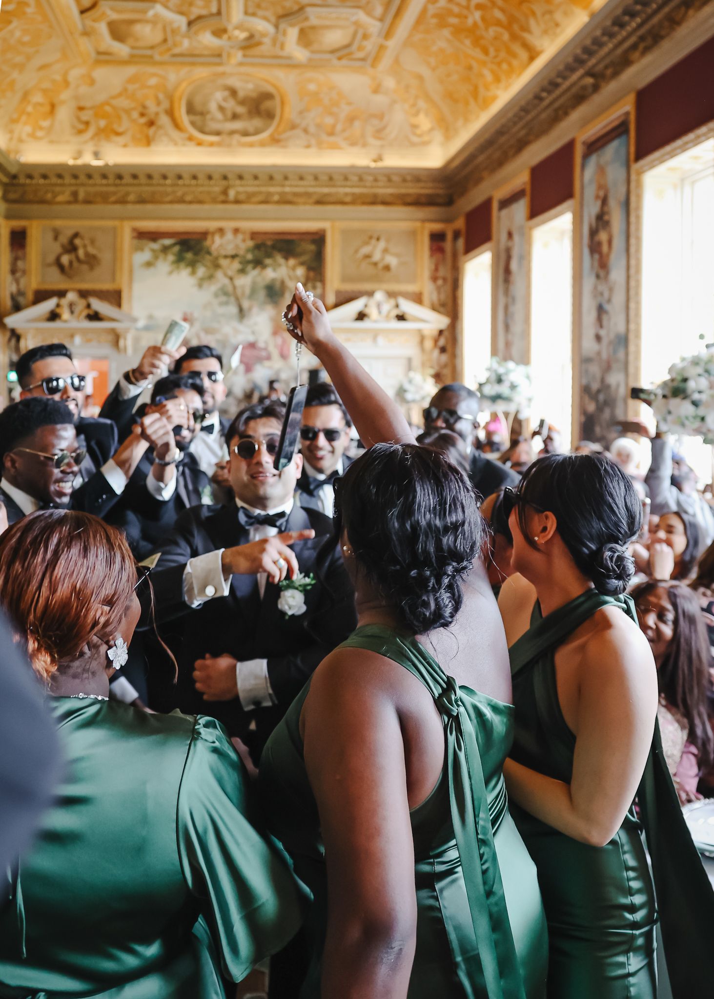 Wedding guests in green dresses and formal attire celebrate in an ornate room with decorative ceiling.