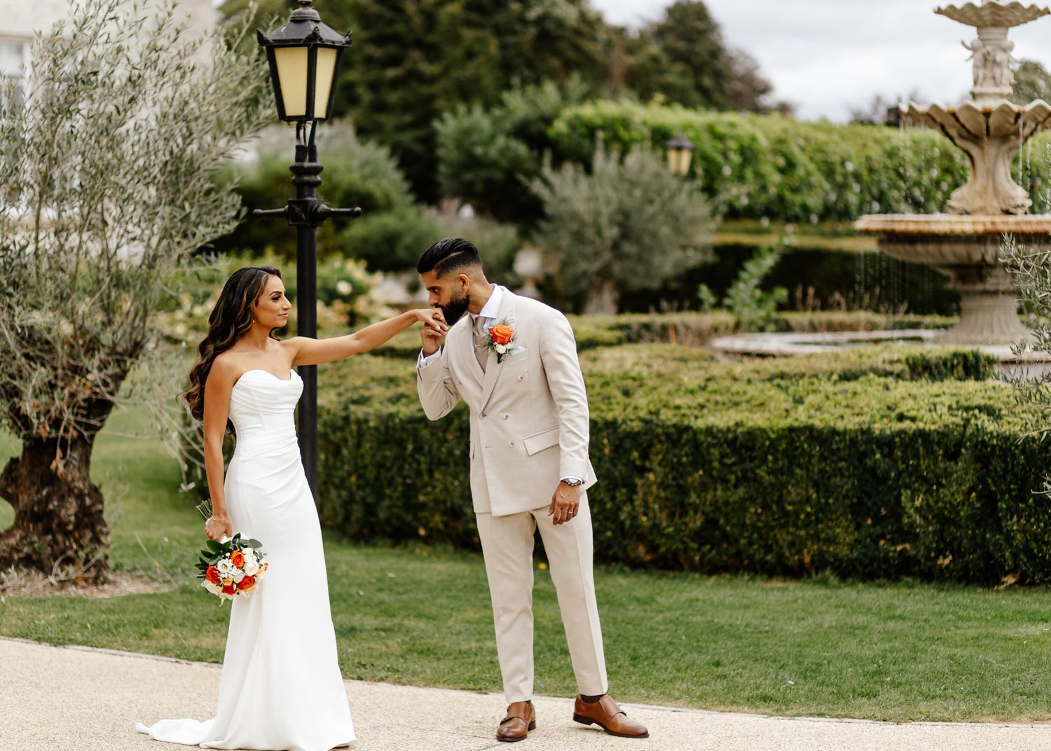 A couple shares a romantic moment in a garden setting with a fountain and lush greenery in the background.