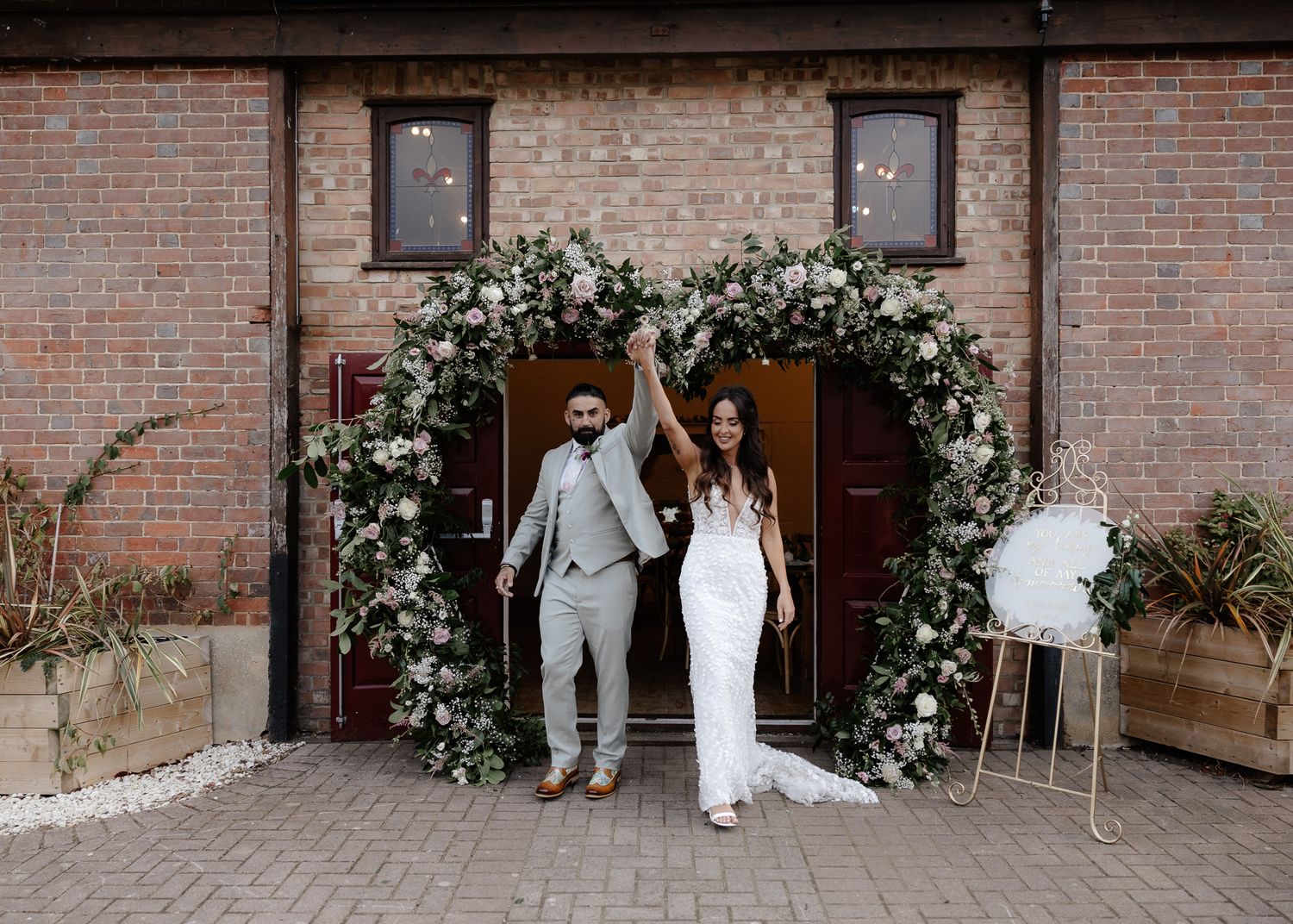 A wedding couple and their guests pose for photos against a rustic brick wall decorated with floral arrangements.