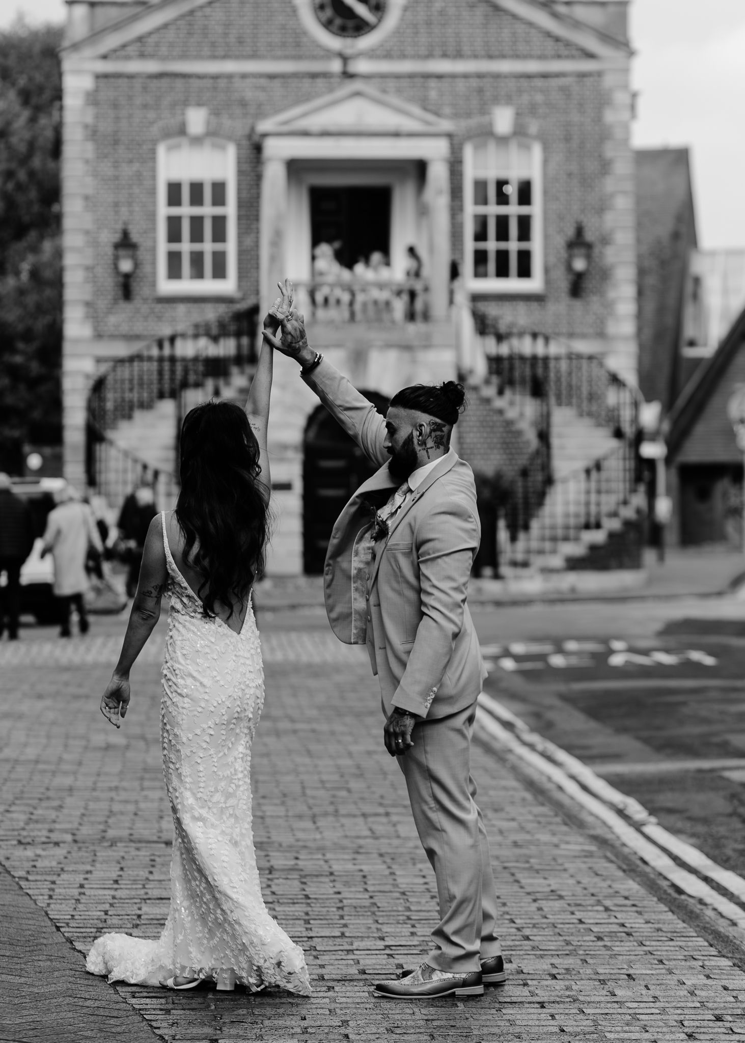 Black and white photo of a couple dancing playfully on cobblestones near a historic building.