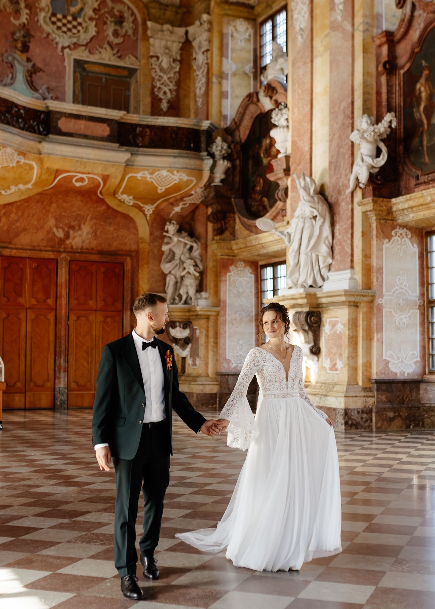 Bride and groom hold hands in an ornate baroque palace hall with marble floors and classical sculptures.