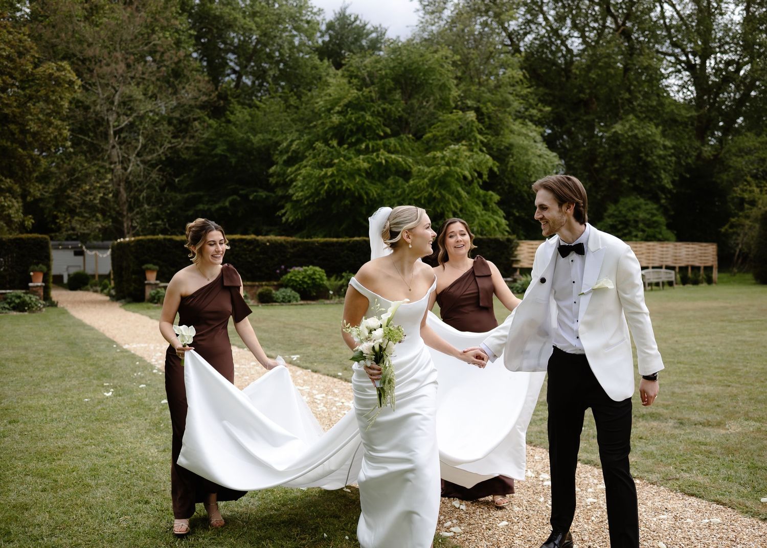 Bride in white gown walks with groom in white tuxedo and bridesmaids in burgundy dresses outdoors.