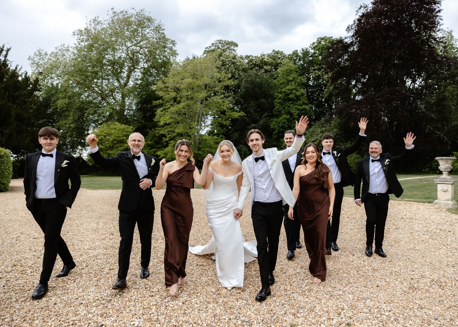 Wedding party celebrating outdoors on gravel path, bride in white gown with groomsmen in tuxedos and bridesmaids in burgundy dresses.
