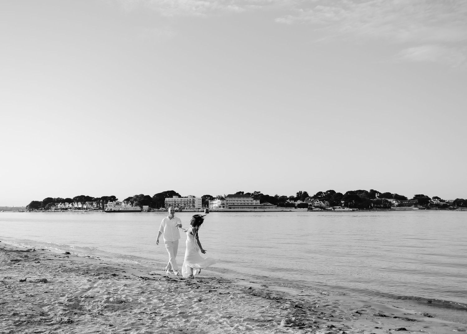 Two people walk hand-in-hand along a sandy beach shoreline with a coastal town visible in the background.