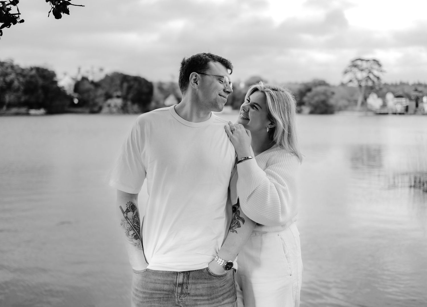 Black and white photo of a couple standing romantically by a lake, the woman leaning close to the man outdoors.