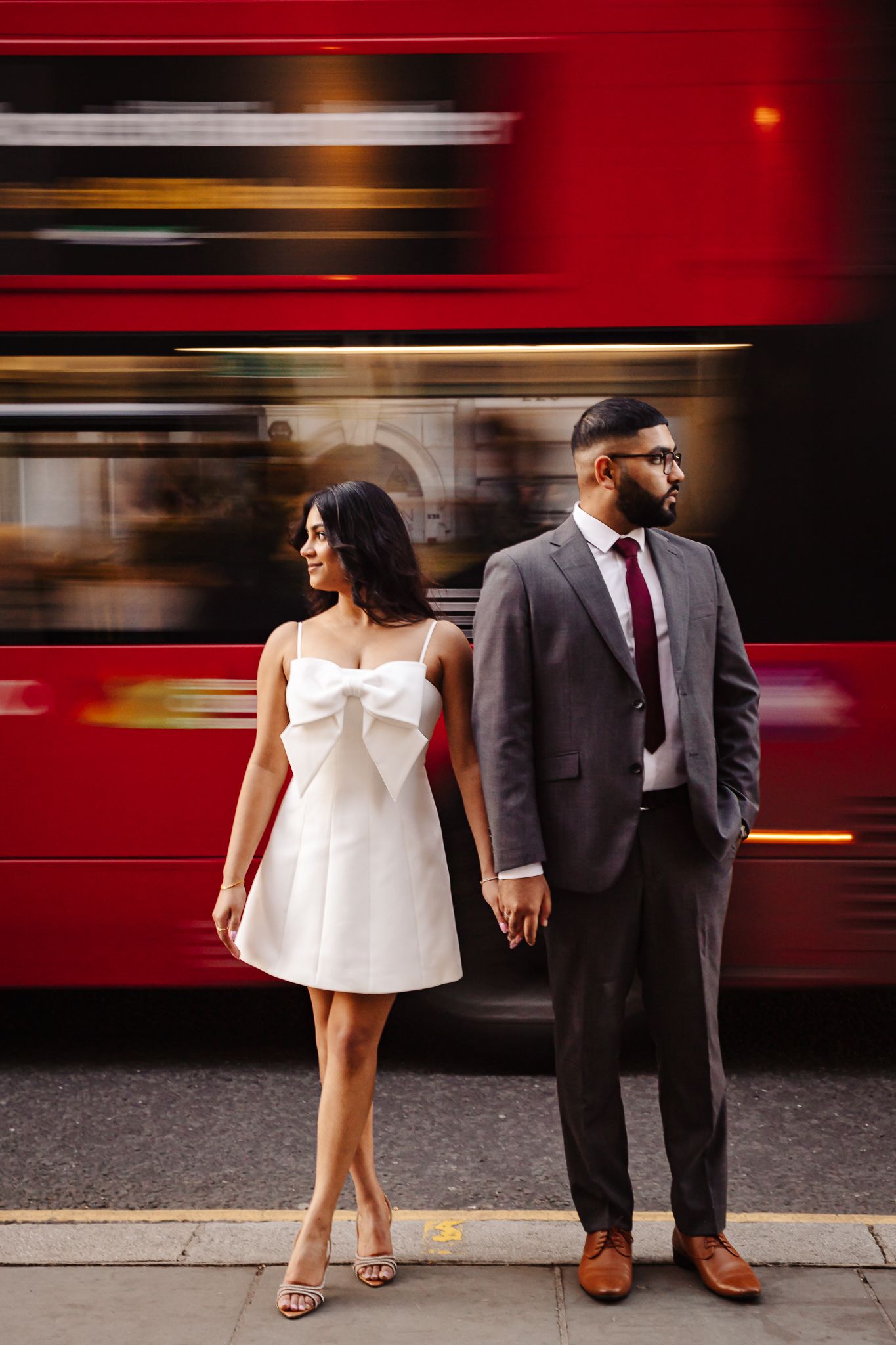 Couple posing on a city sidewalk with a blurred red double-decker bus passing behind them.