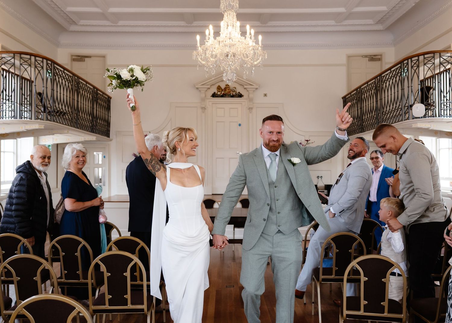 Joyful newlyweds celebrate walking down the aisle in an elegant ballroom with chandelier, guests cheering around them.