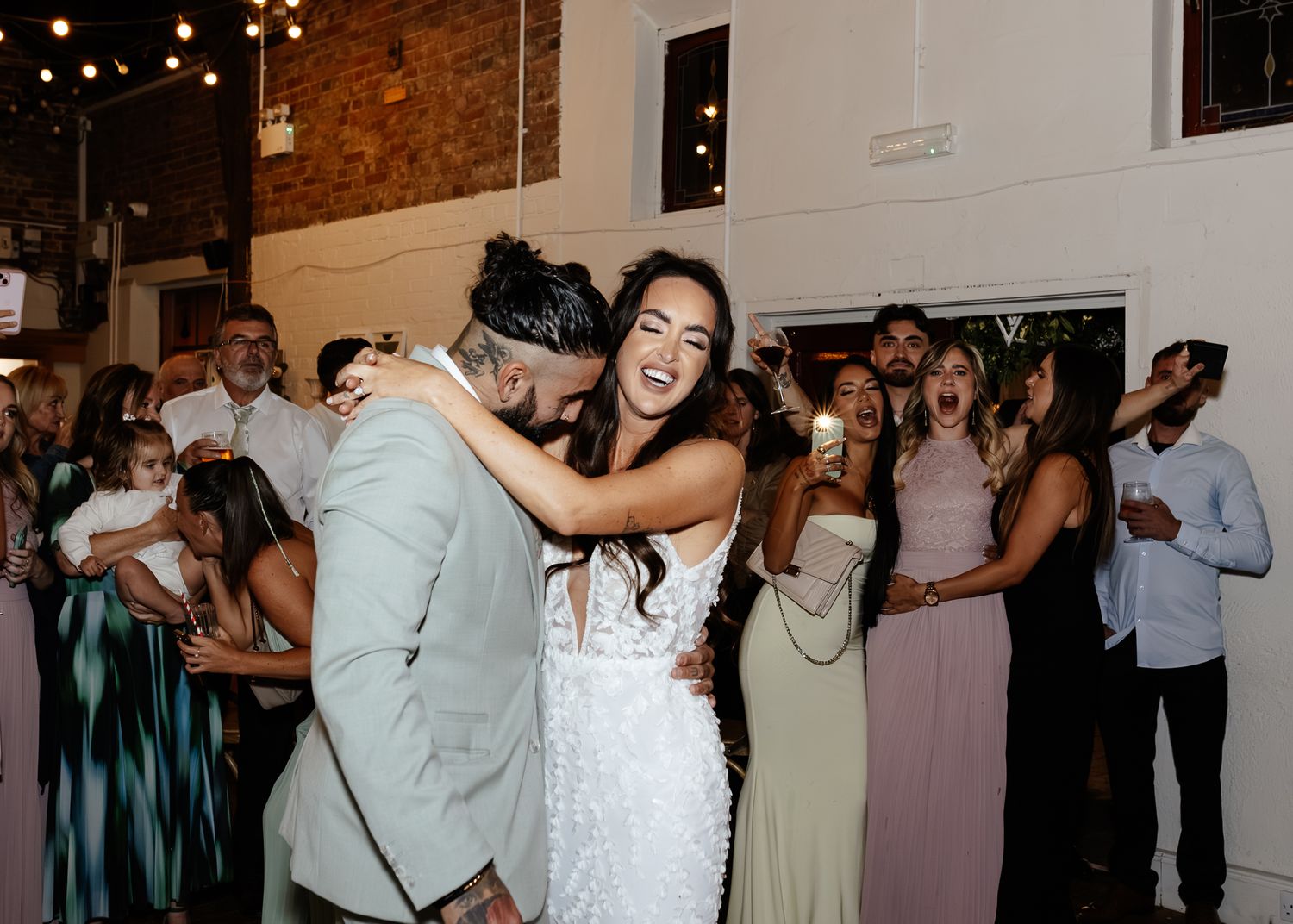 Wedding guests dance and celebrate at a reception with string lights and brick walls in the background.