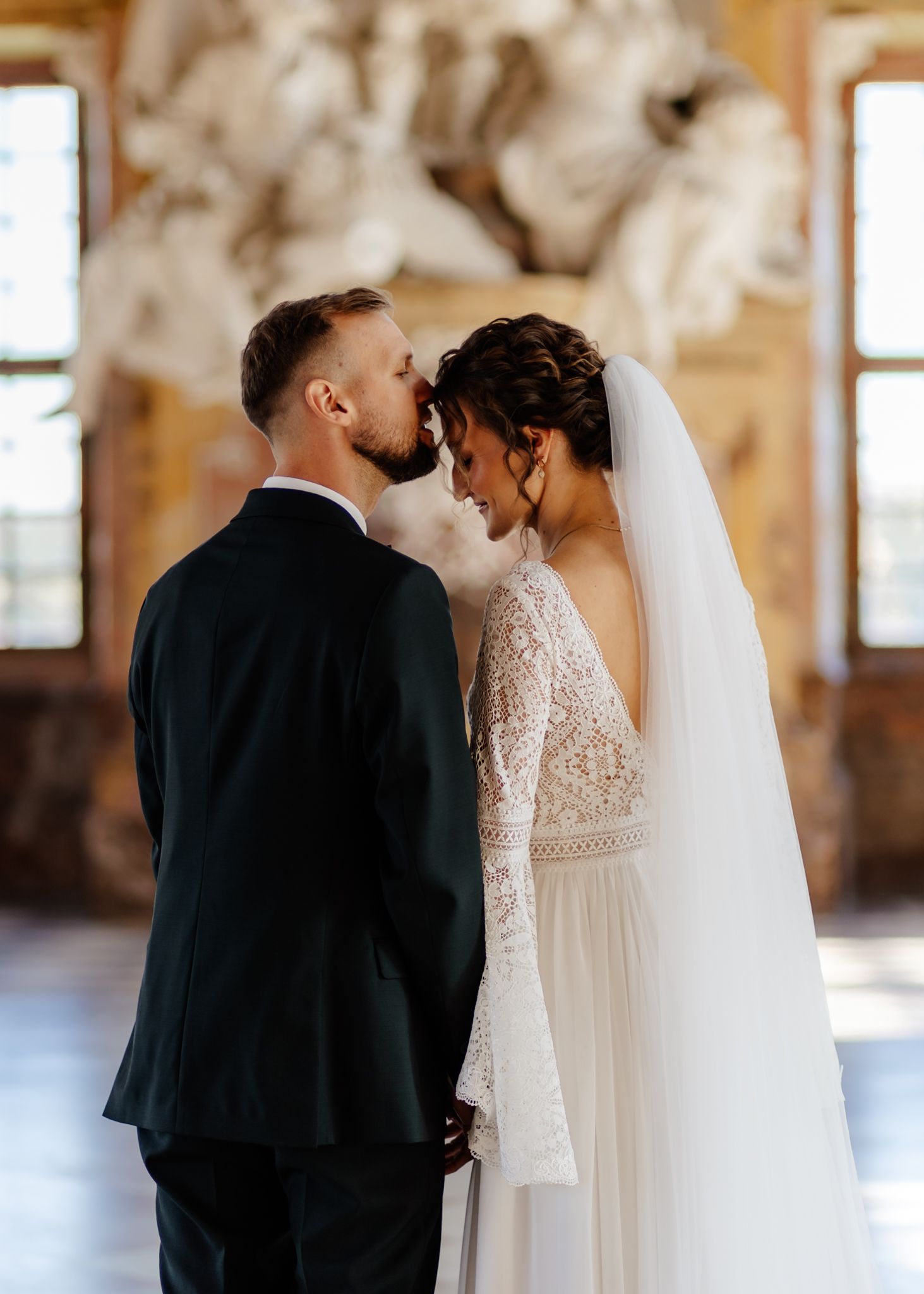 Newlyweds share an intimate moment in a grand hall with sunlit windows and classical architecture.