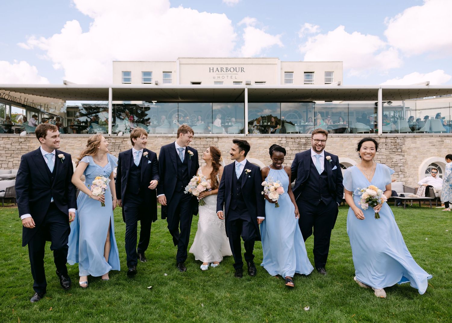 Wedding party in navy suits and blue gowns walks on lawn outside Harbour venue on a sunny day.