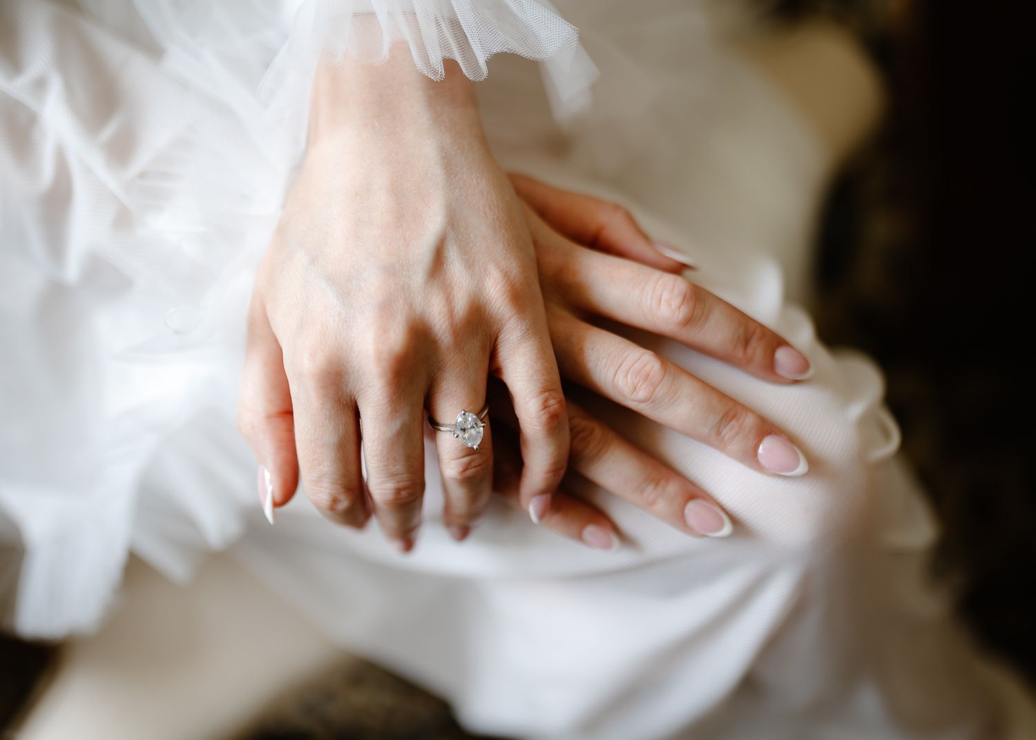 Close-up details of a bride's hands wearing a white wedding dress with delicate lace and a sparkling engagement ring.