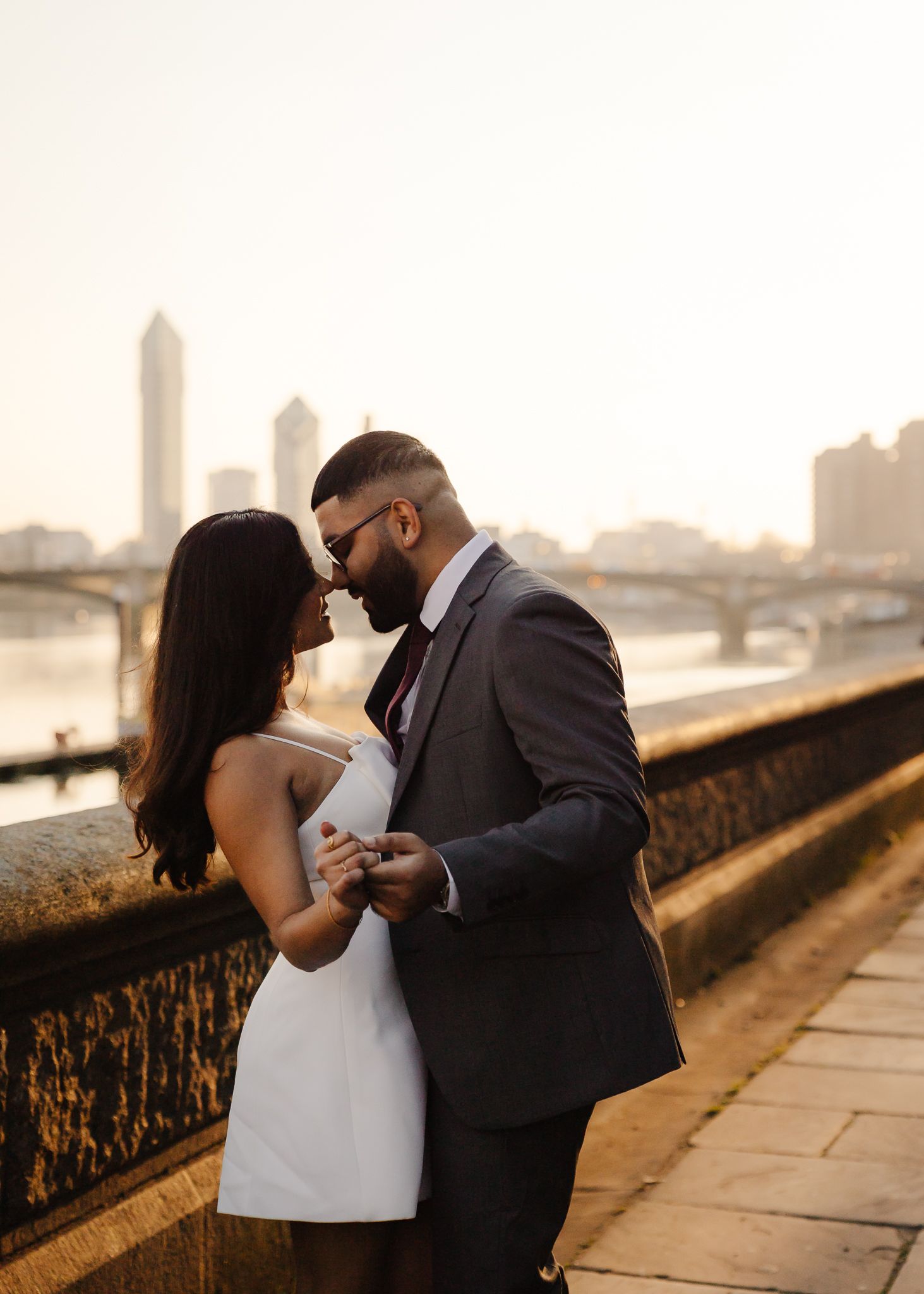 A romantic couple shares intimate moments along a stone wall during sunset in an urban setting.