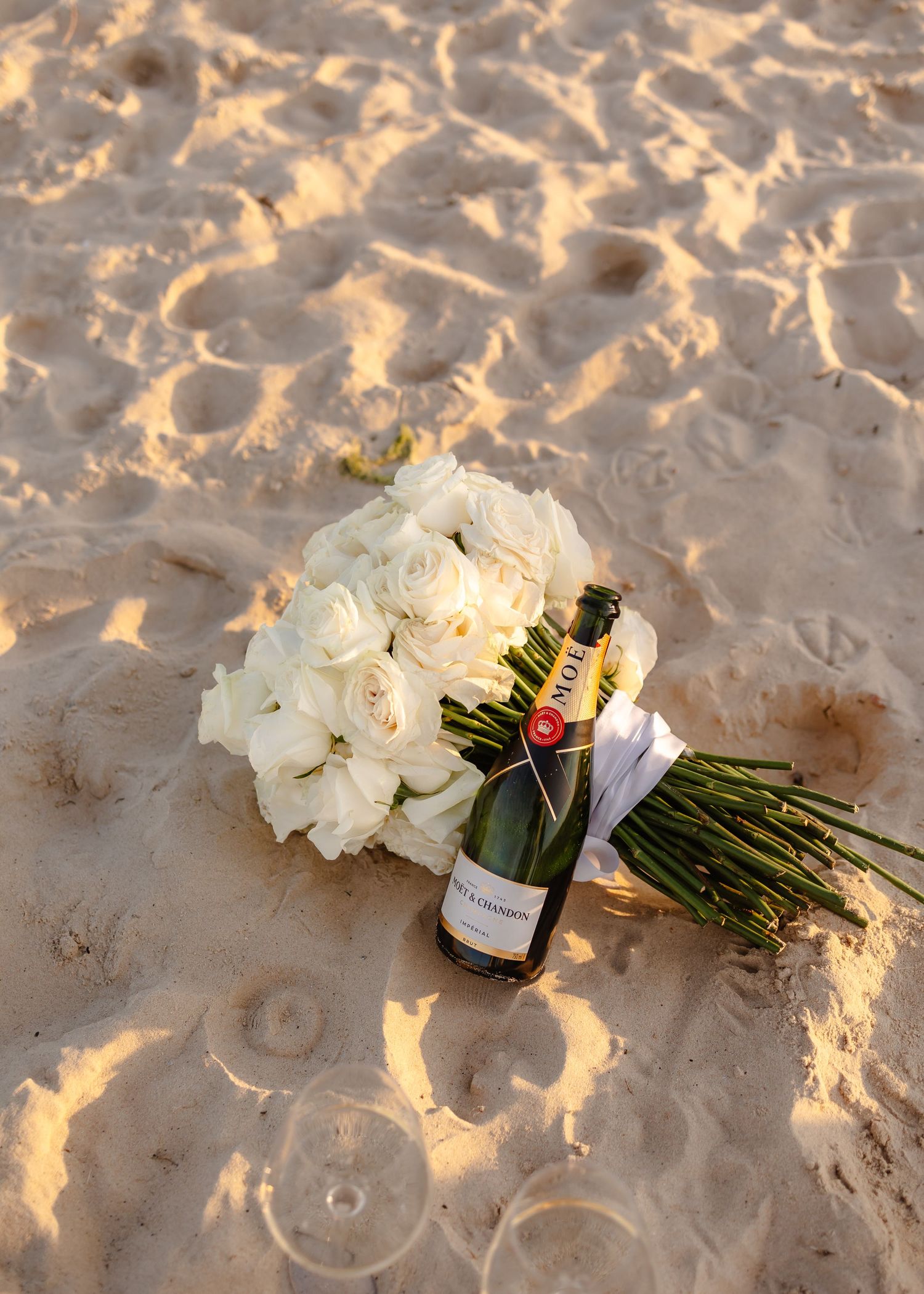 White rose bouquet and Moet champagne bottle resting on sandy beach, perfect for weddings or celebrations.