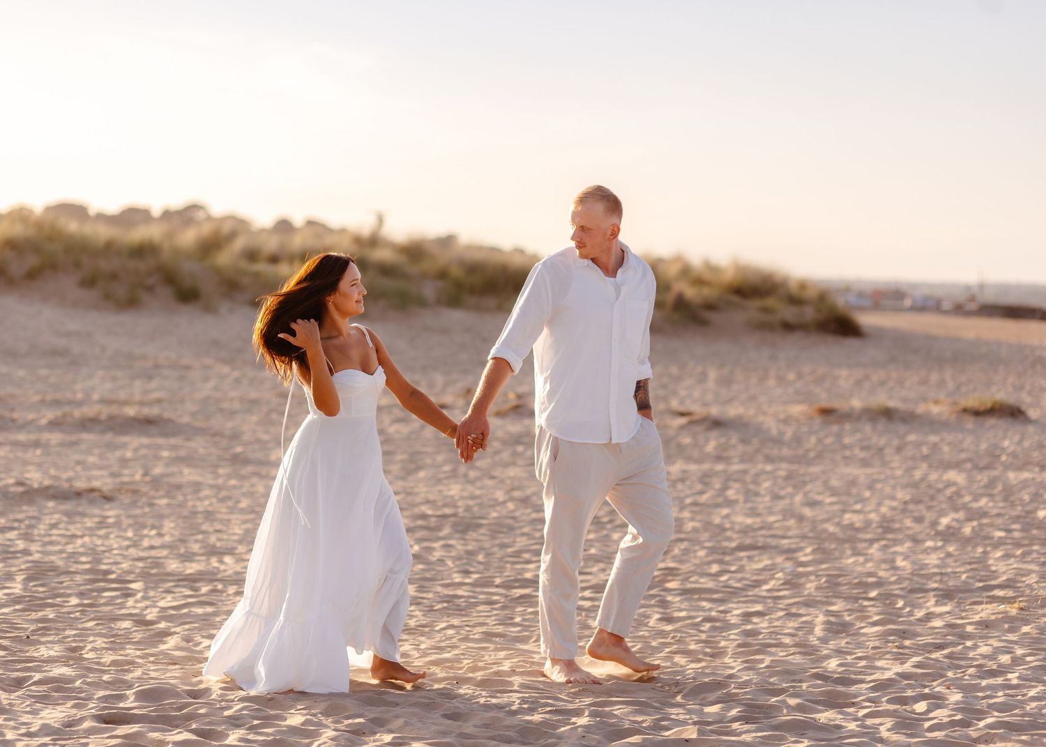Couple in white outfits holding hands walking barefoot on sandy beach at golden sunset.