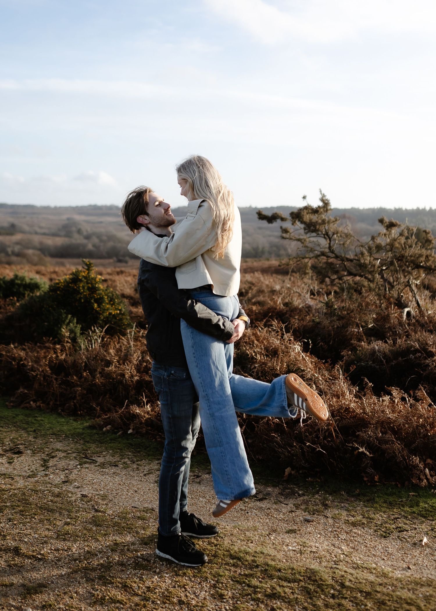 A man lifts a smiling blonde woman outdoors in a scenic heathland landscape under a bright sky.