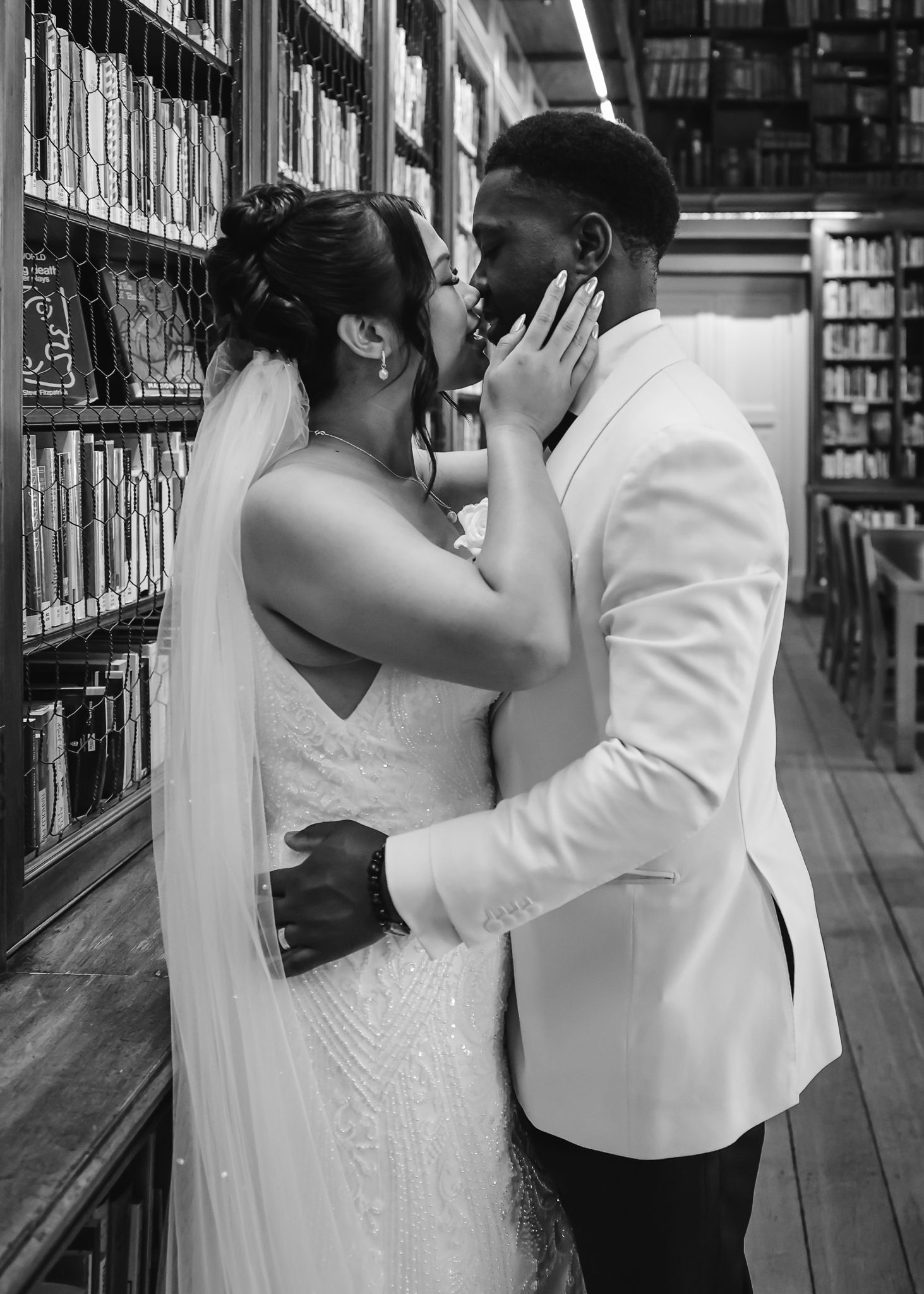A romantic wedding photo of a couple sharing an intimate moment between library bookshelves in black and white.