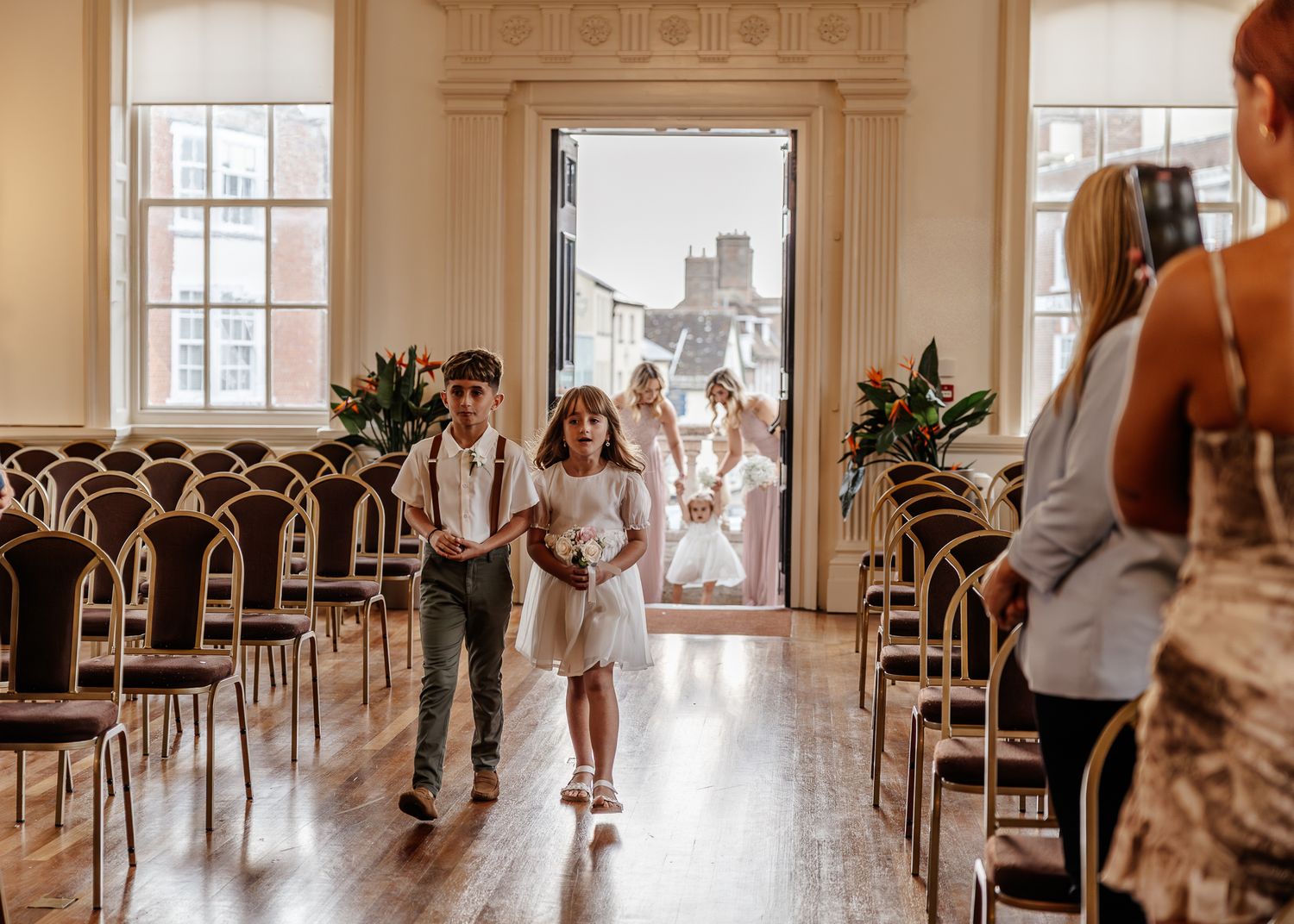 Wedding guests mingle and socialize in an elegant white ballroom with hardwood floors and large windows.